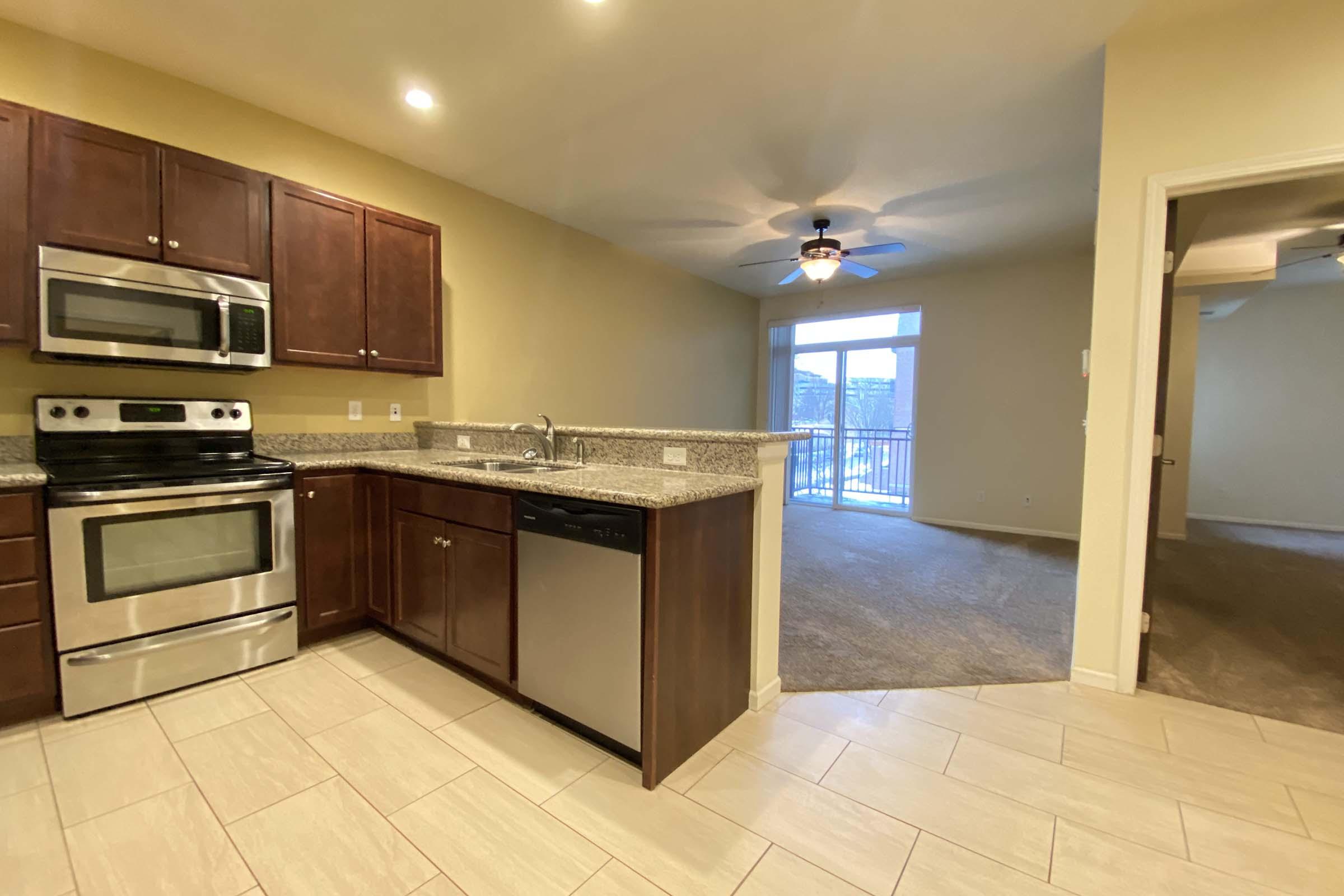 Modern kitchen featuring dark wood cabinets, stainless steel appliances, including a microwave and dishwasher, and granite countertops. There's a view of a spacious living area with a ceiling fan and sliding glass doors leading to a balcony or patio. Light-colored tile flooring throughout.