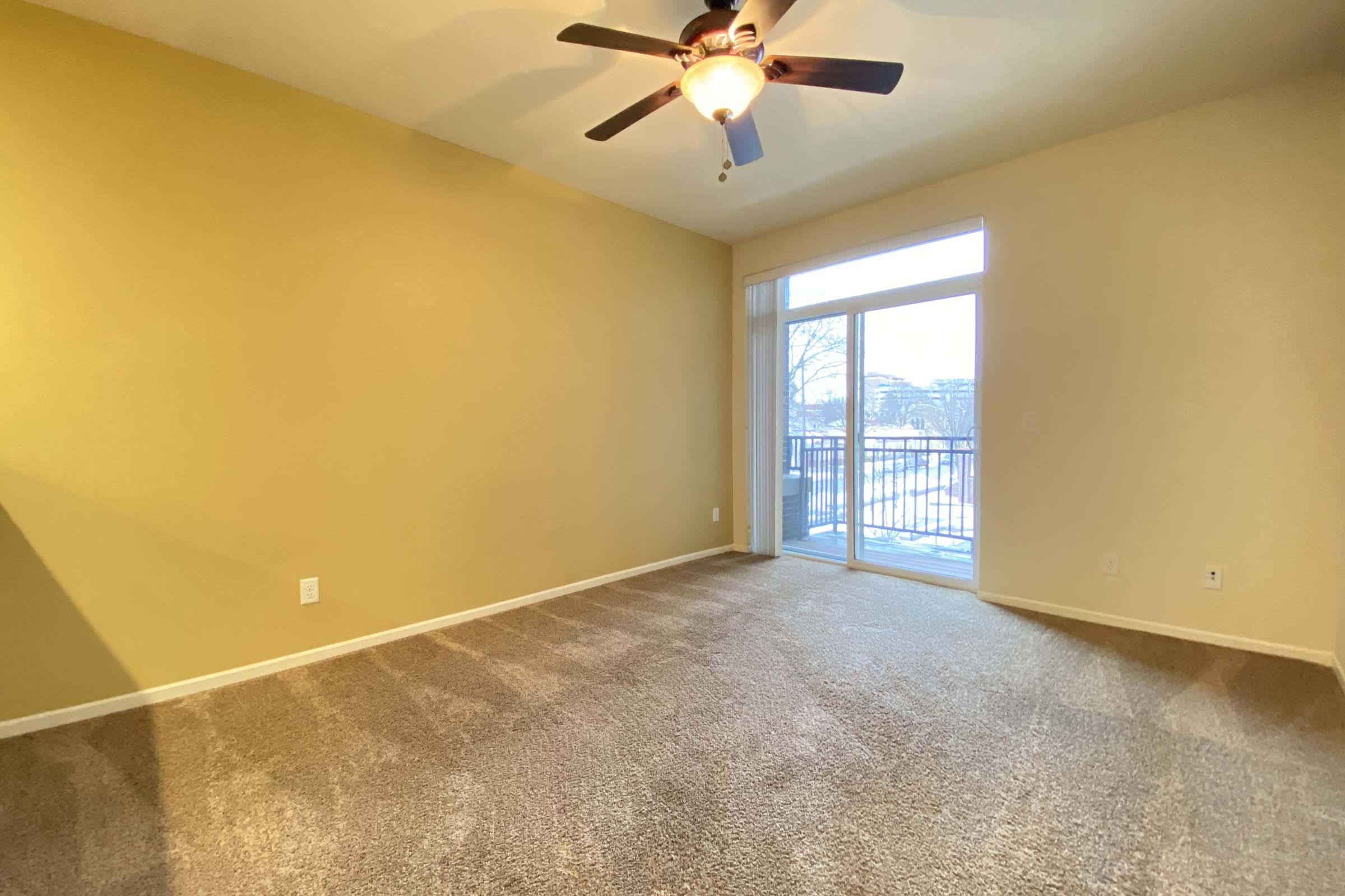 A spacious, empty room with beige walls and a light brown carpet. A ceiling fan is installed above, and there is a large window with sliding doors leading to a balcony. Natural light streams into the room, illuminating the space, which has no furniture.