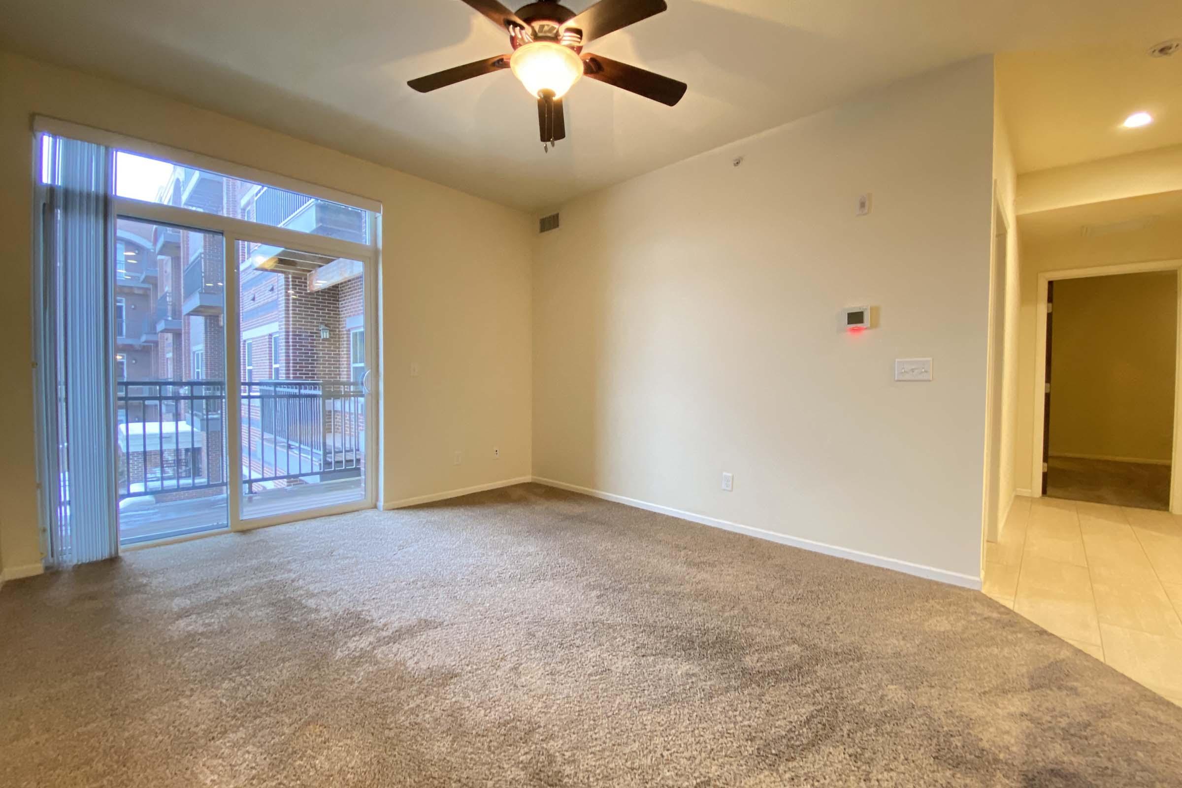Spacious living room with a ceiling fan, beige carpet, and neutral-colored walls. A sliding glass door leads to a small balcony, offering natural light. The room features a corner with a thermostat, and an open entryway to another room, creating a warm, inviting atmosphere.