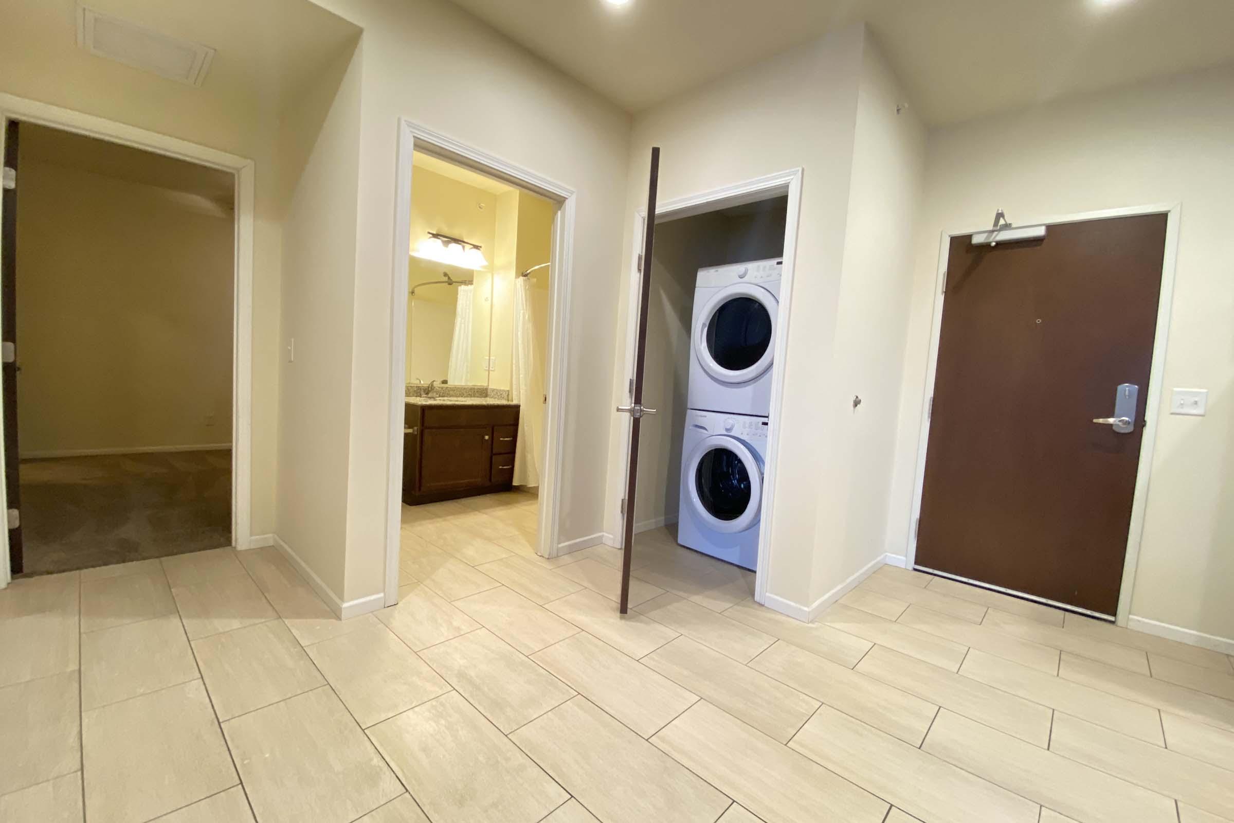 A spacious interior hallway featuring two open doorways leading to separate rooms. One doorway opens to a bathroom with a vanity, and the other leads to an empty room. A stacked washer and dryer unit is positioned along the wall, and the entrance door is visible on the right. The floors are tiled and well-lit.