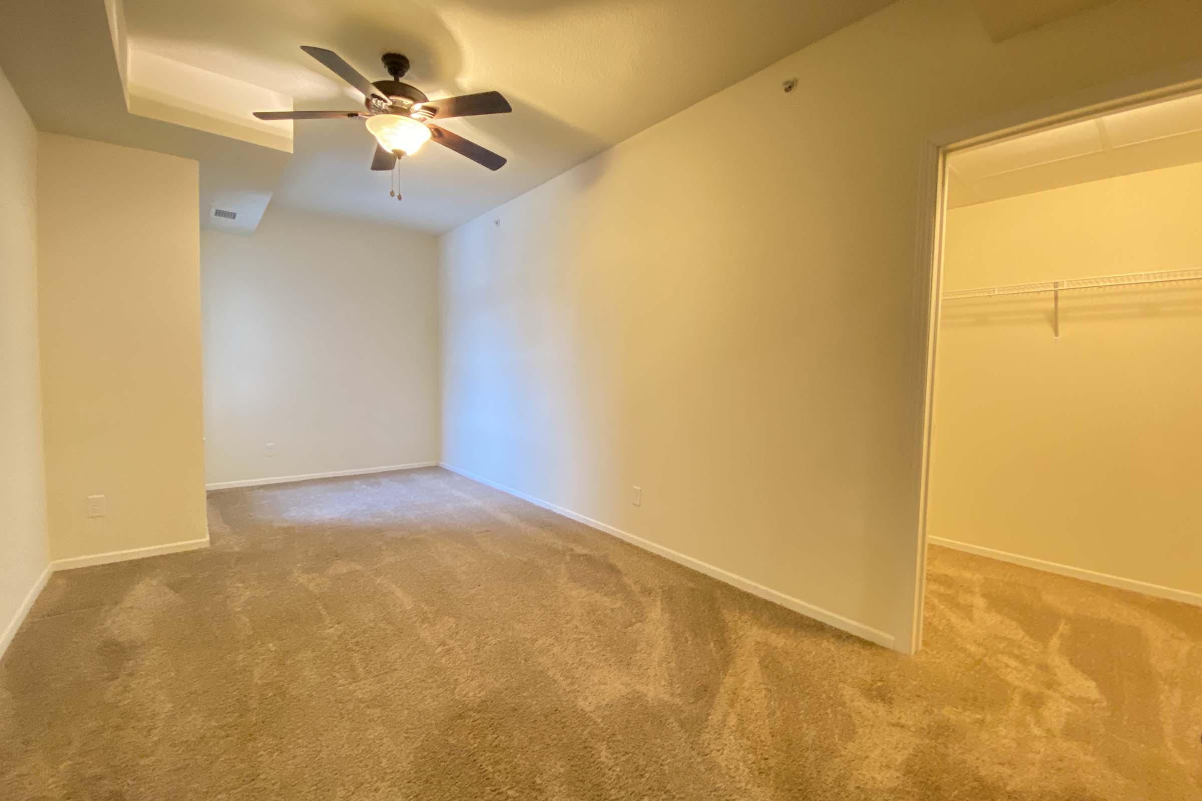 A spacious, well-lit room with beige carpet and light yellow walls. It features a ceiling fan with light, a closet with a sliding door, and a large window allowing natural light. The room is empty, creating a blank canvas for decoration.