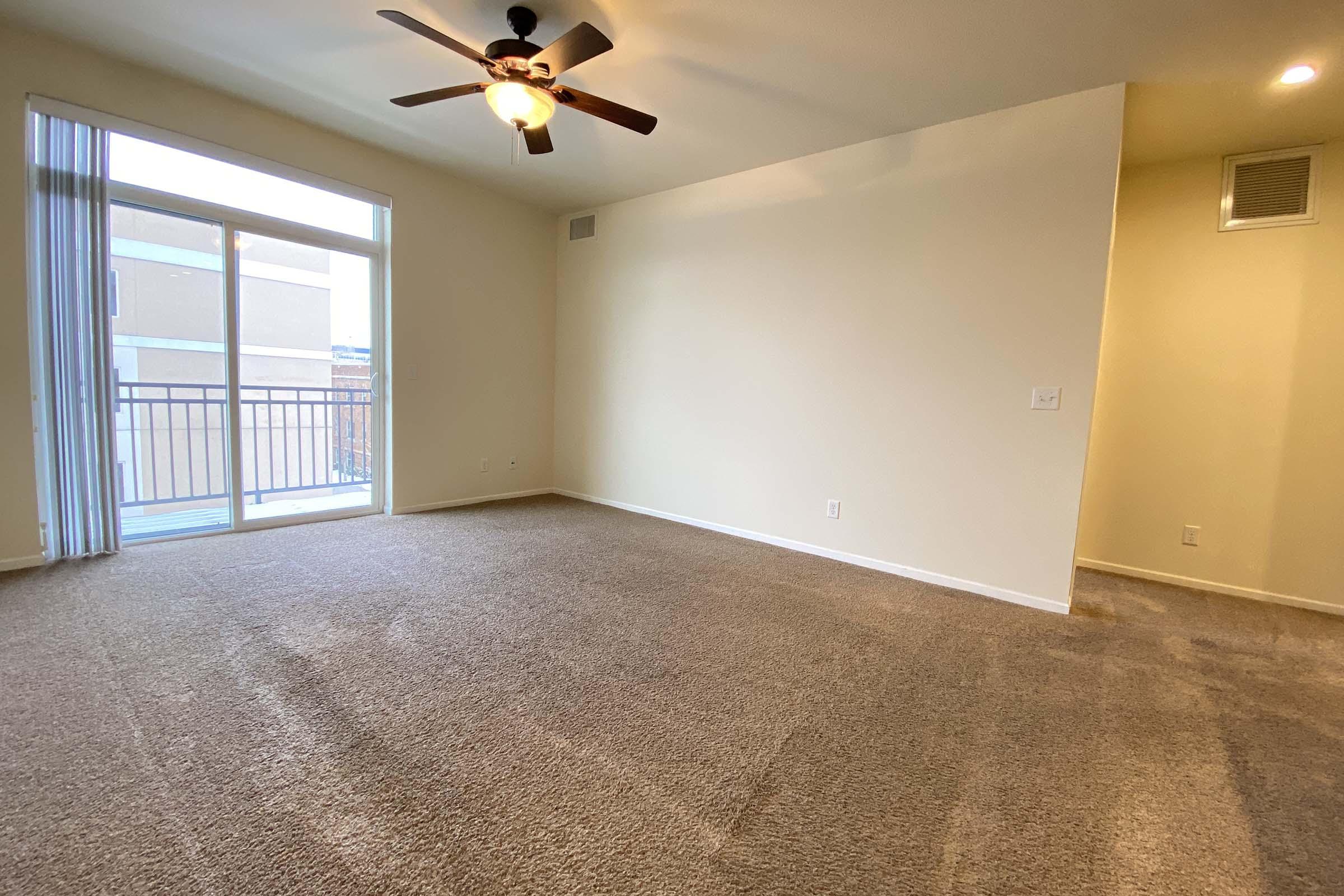 A spacious, empty room with light beige walls and a ceiling fan. Large sliding glass doors lead to a balcony, providing natural light. The floor is covered in light brown carpet, creating a cozy atmosphere. One corner features a small wall with an air vent.