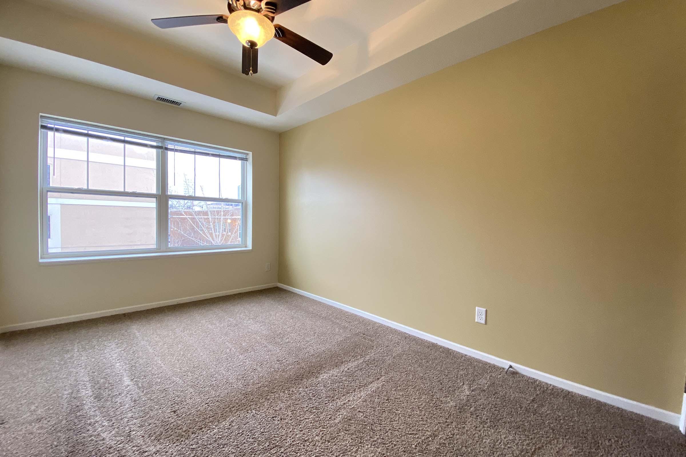 Empty room with beige walls and a ceiling fan. The floor is covered in light-colored carpet, and there are large windows allowing natural light to enter. The overall atmosphere is bright and spacious, with no furniture present.