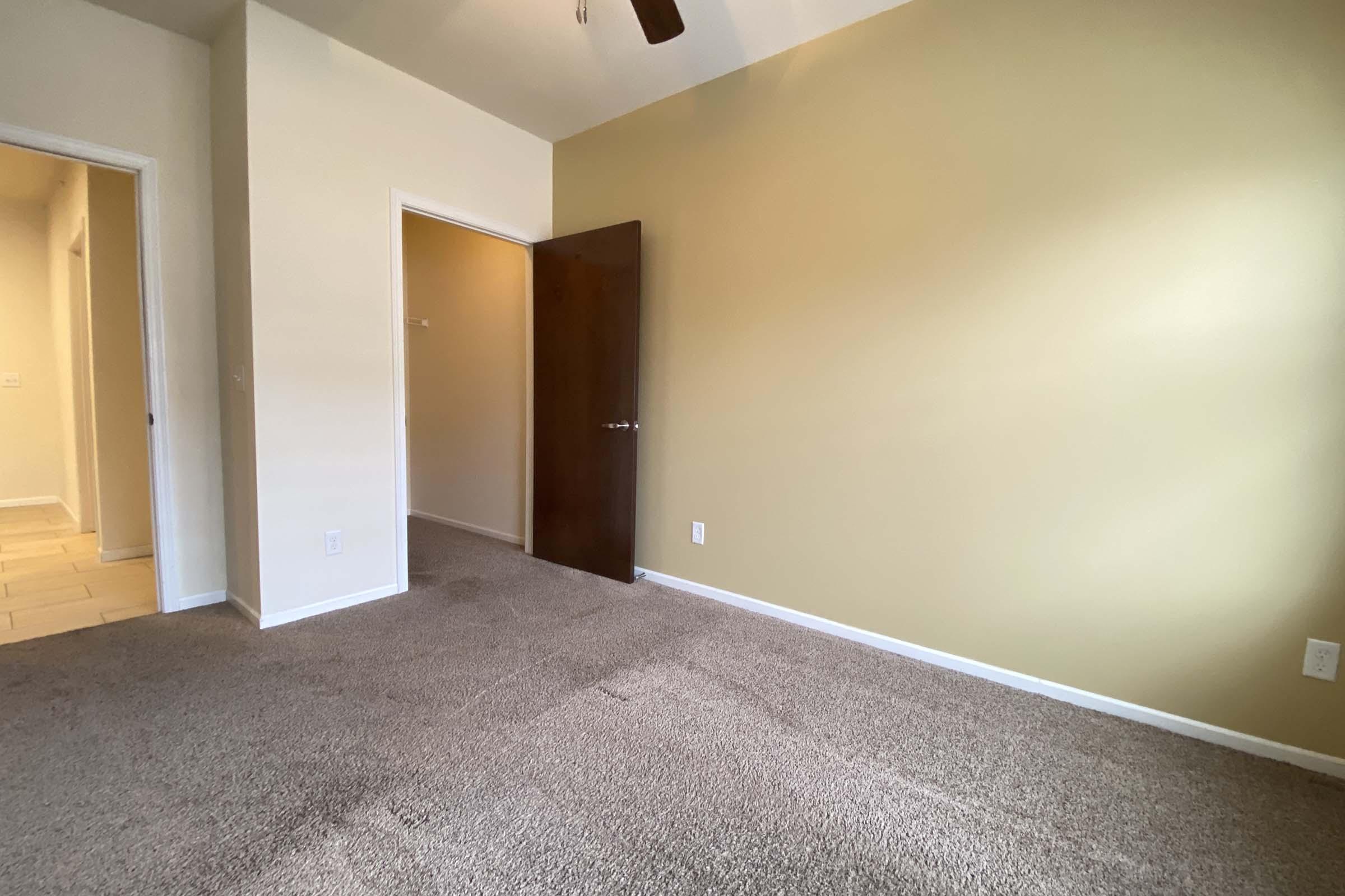 A well-lit room with beige walls and carpeted flooring. There is a closed wooden door on the right leading to another space, and a doorway on the left leading to a hallway. The room appears empty and neatly maintained, with natural light coming in from an unseen window.