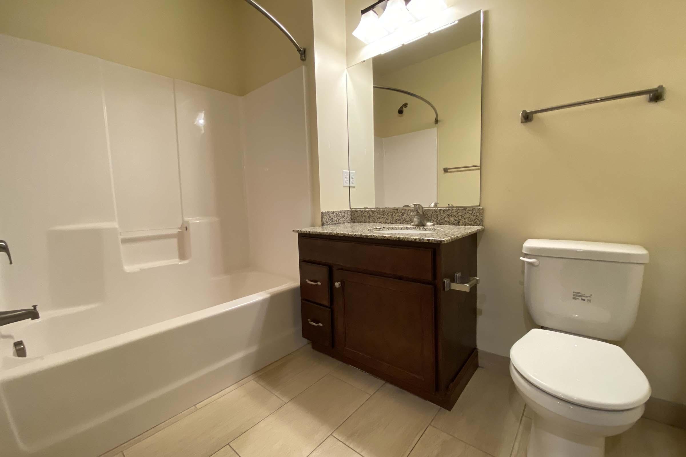 A clean, modern bathroom featuring a bathtub with shower, a granite countertop vanity with a sink, a large mirror, and a white toilet. The walls are painted a light color and the flooring consists of neutral tiles. The space is well-lit with overhead light fixtures.