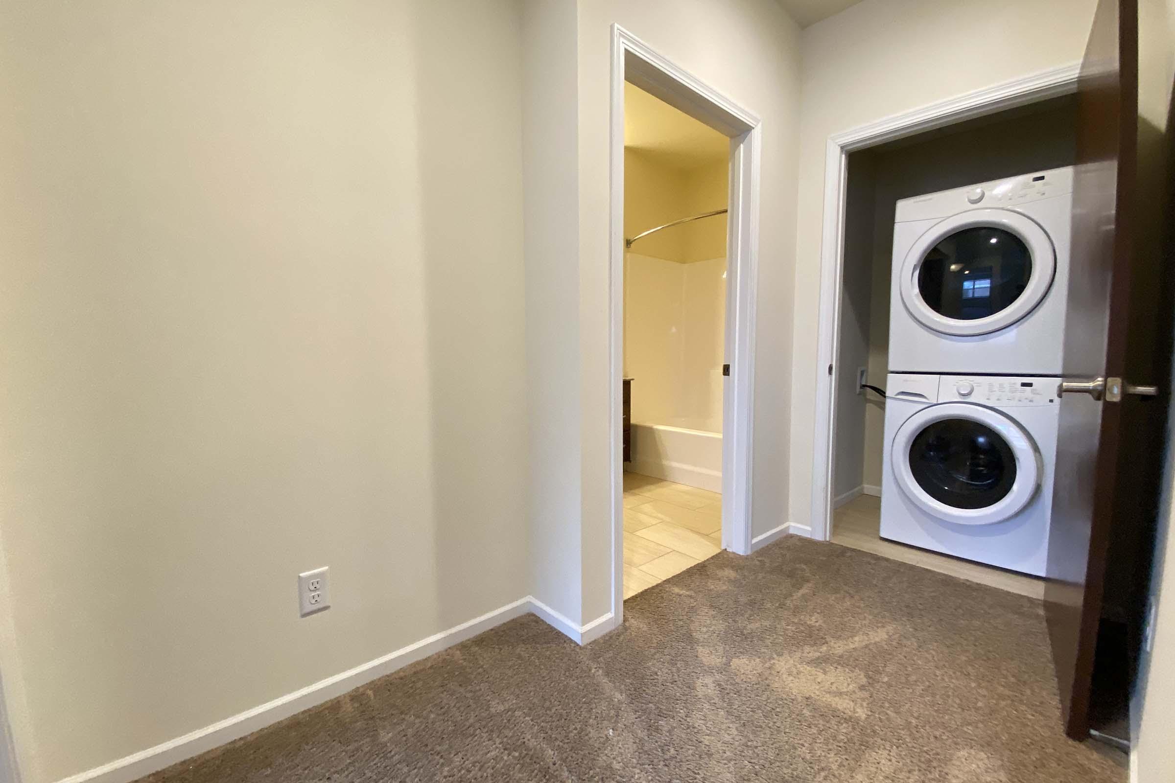 View of a laundry area featuring a stacked washer and dryer against a light-colored wall. A doorway leads to a bathroom on the left, with bright lighting suggesting it's night. The floor is carpeted in a neutral tone, and the space appears clean and organized.