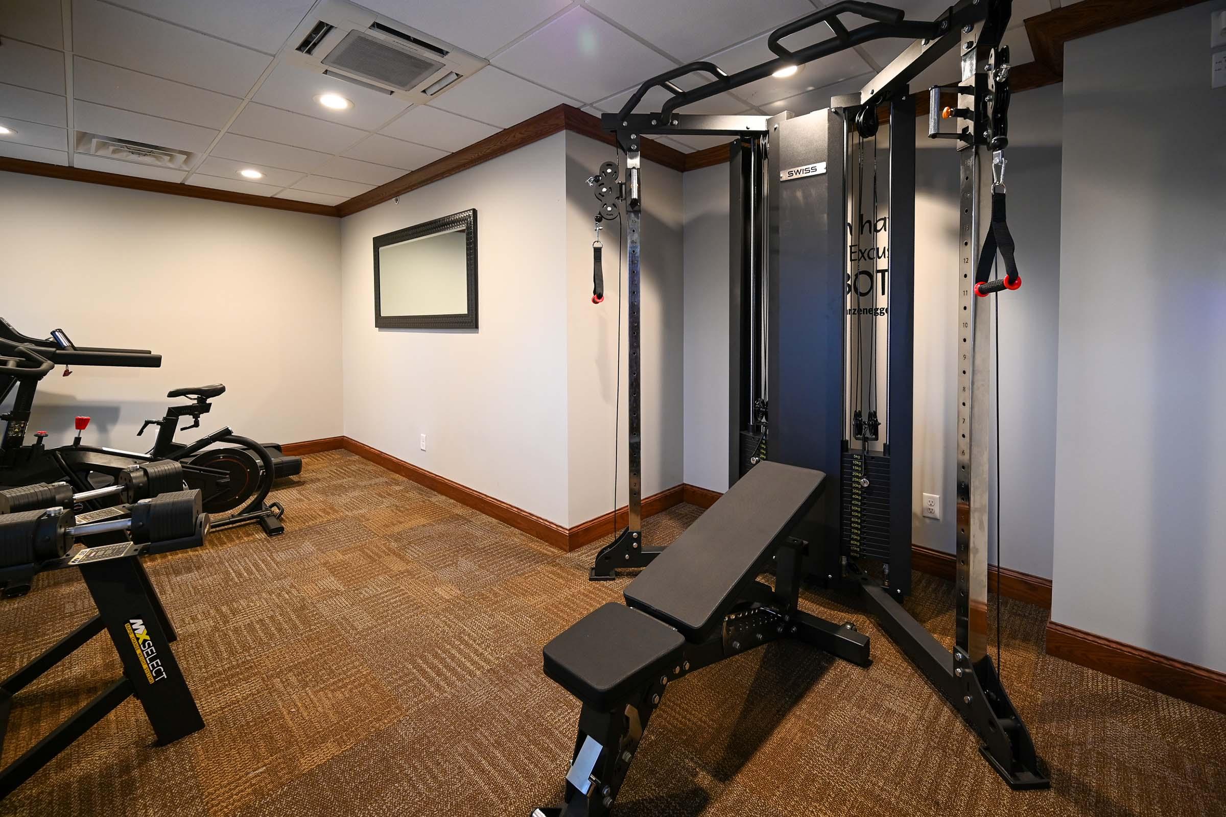 A well-equipped home gym featuring a multi-functional weight machine with adjustable cables, a flat workout bench, and various exercise machines in a cozy interior space with neutral walls and carpeted flooring.