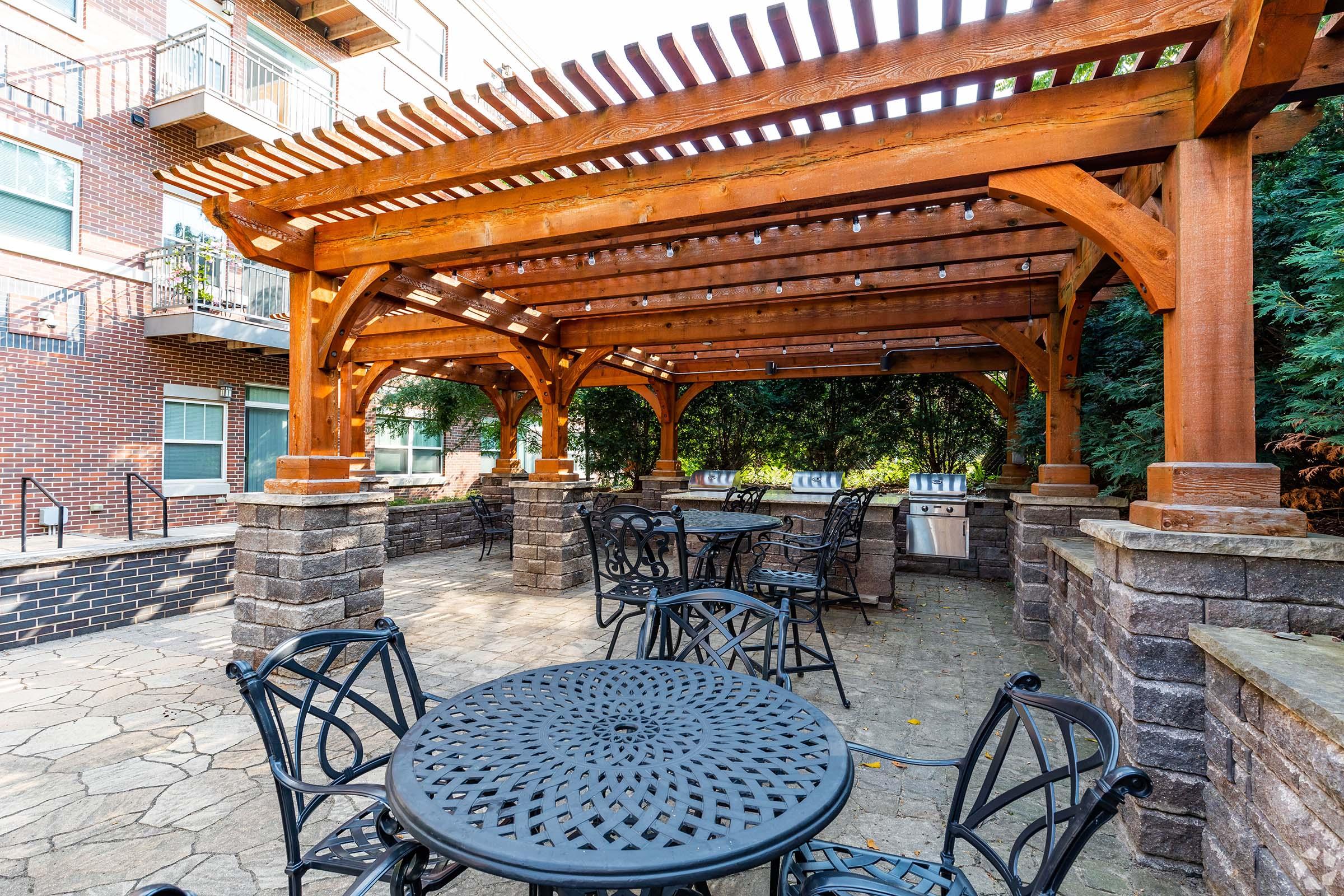 A landscaped outdoor area featuring a wooden pergola with a slatted roof, surrounded by stone columns. Below, there are several black wrought-iron tables and chairs on a stone patio. In the background, there are gas grills and lush greenery, creating a welcoming space for gatherings.