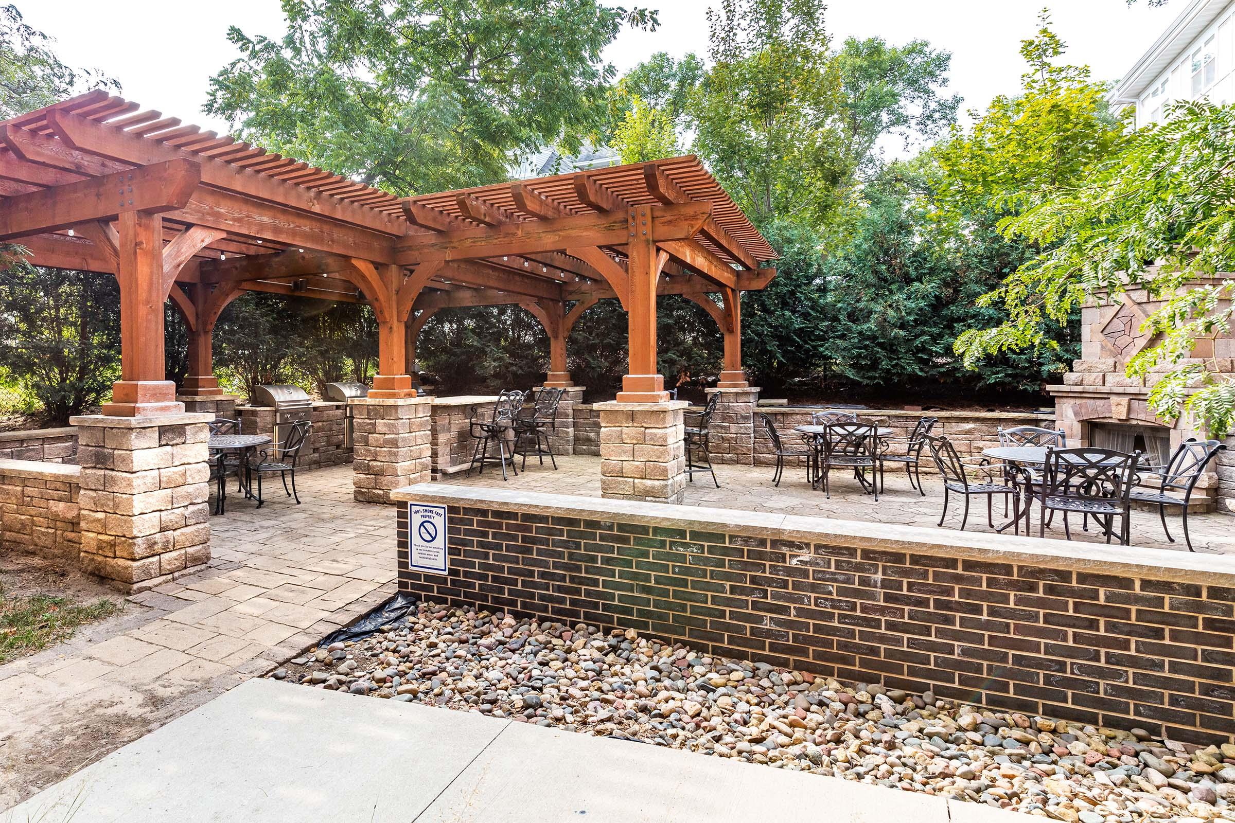 A landscaped outdoor space featuring a wooden pergola, several tables and chairs, and a stone wall. Lush greenery surrounds the area, providing a serene atmosphere for visitors. The ground is paved with stone and complemented by decorative gravel.