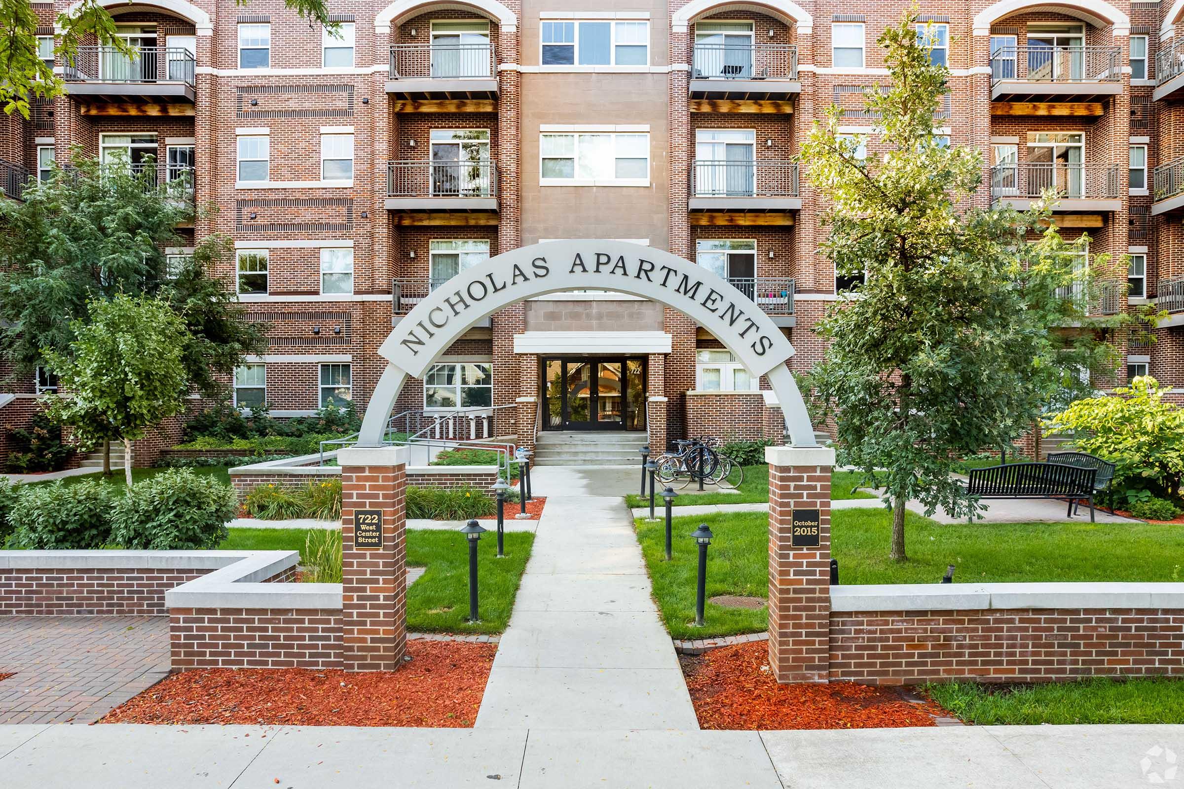 Archway labeled "Nicholas Apartments" leading to a multi-story brick apartment building. The entrance is flanked by trees and landscaped areas. A pathway runs through the well-maintained grounds, with a bicycle rack and benches visible in the area.