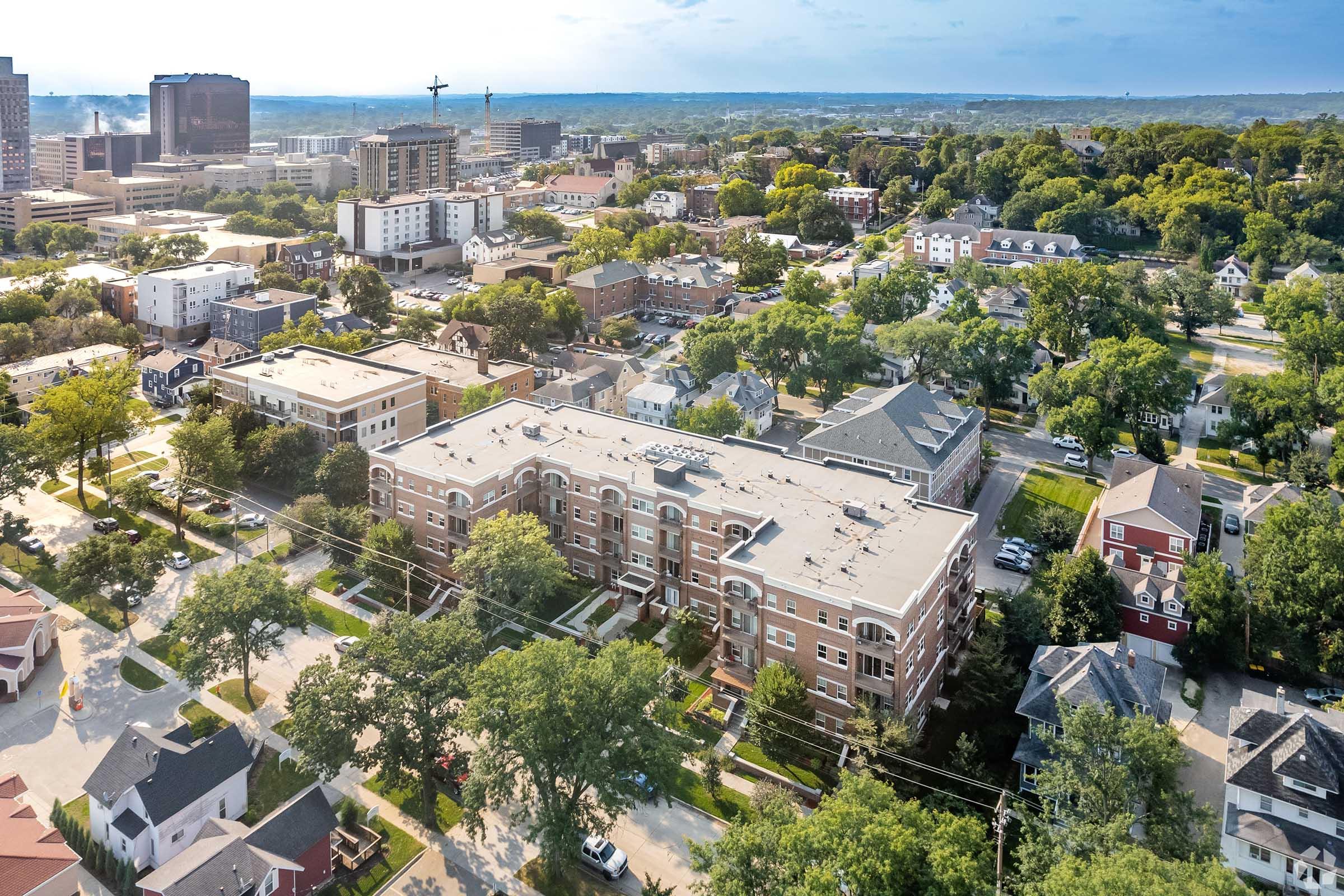 Aerial view of a diverse urban area featuring residential buildings and greenery, with a mix of multi-story apartments and single-family homes. In the background, city skyscrapers and distant hills are visible under a cloudy sky. Streets are lined with trees, showcasing a blend of city living and nature.
