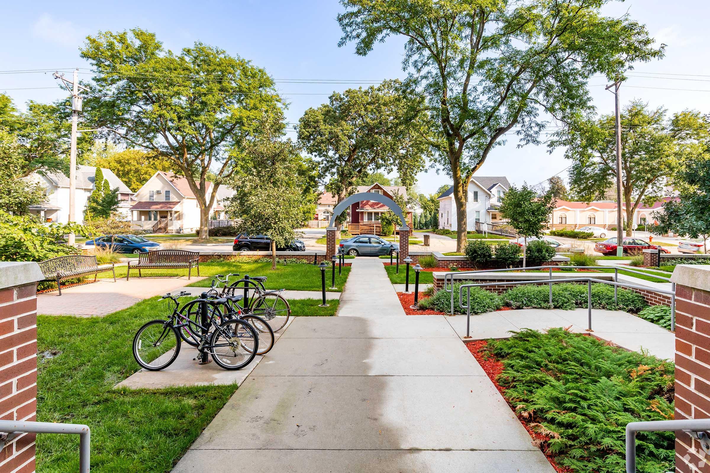 A view from a building entrance showcasing a pathway leading through a park-like area. The path is bordered by green grass and landscaped flower beds. Bicycles are parked on the left, and residential homes are visible in the background under a clear blue sky. Trees provide shade along the path.
