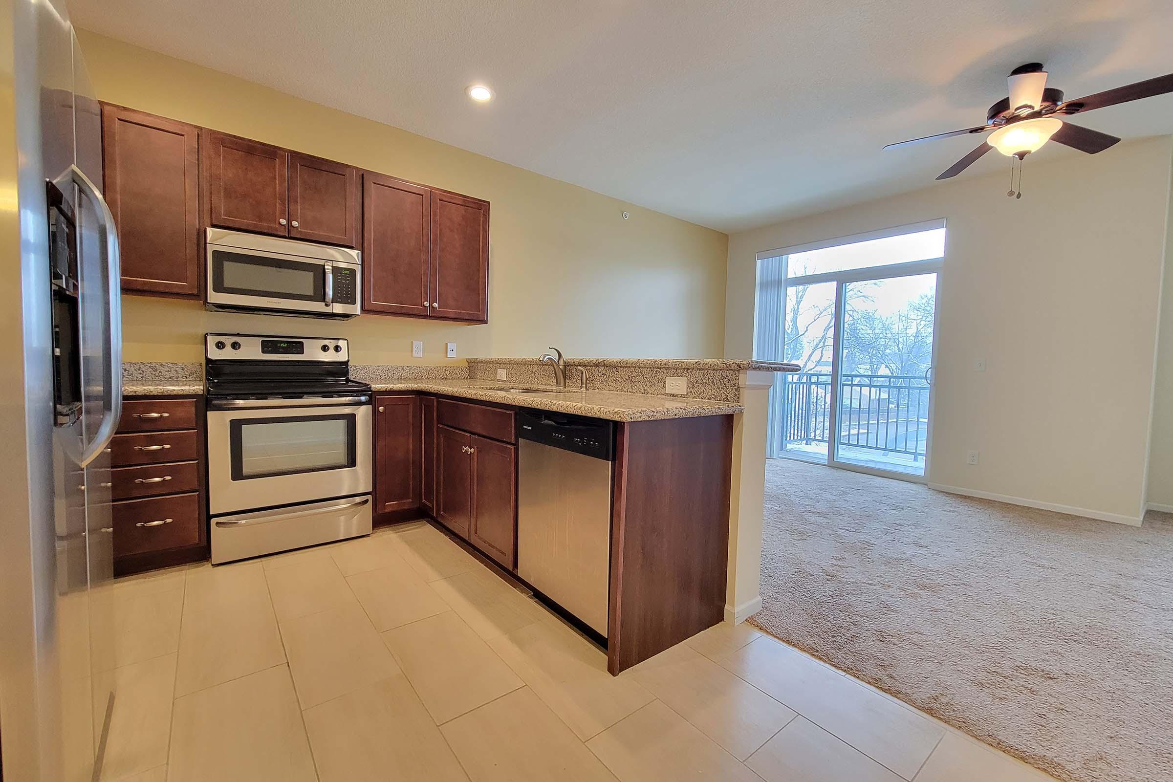 Modern kitchen with stainless steel appliances, including a refrigerator, oven, and dishwasher. Dark wood cabinetry and granite countertops are featured, along with light-colored tile flooring. A spacious living area with carpet is visible in the background, leading to a large window with natural light coming in.