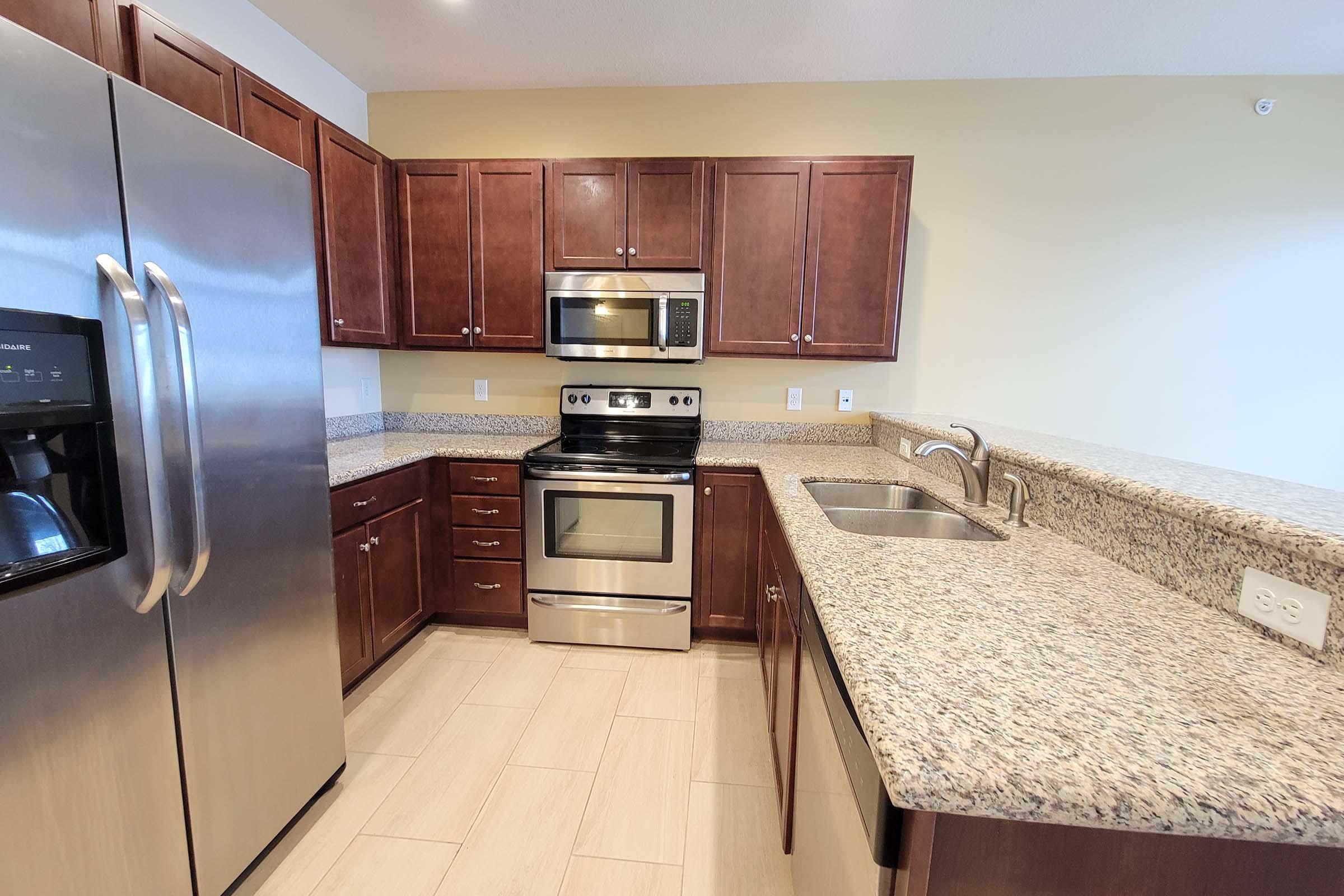 Modern kitchen featuring dark wood cabinets, stainless steel appliances including a refrigerator, oven, and microwave. The countertop is made of granite with a light-colored tile floor. The kitchen has an open layout and large amounts of natural light.