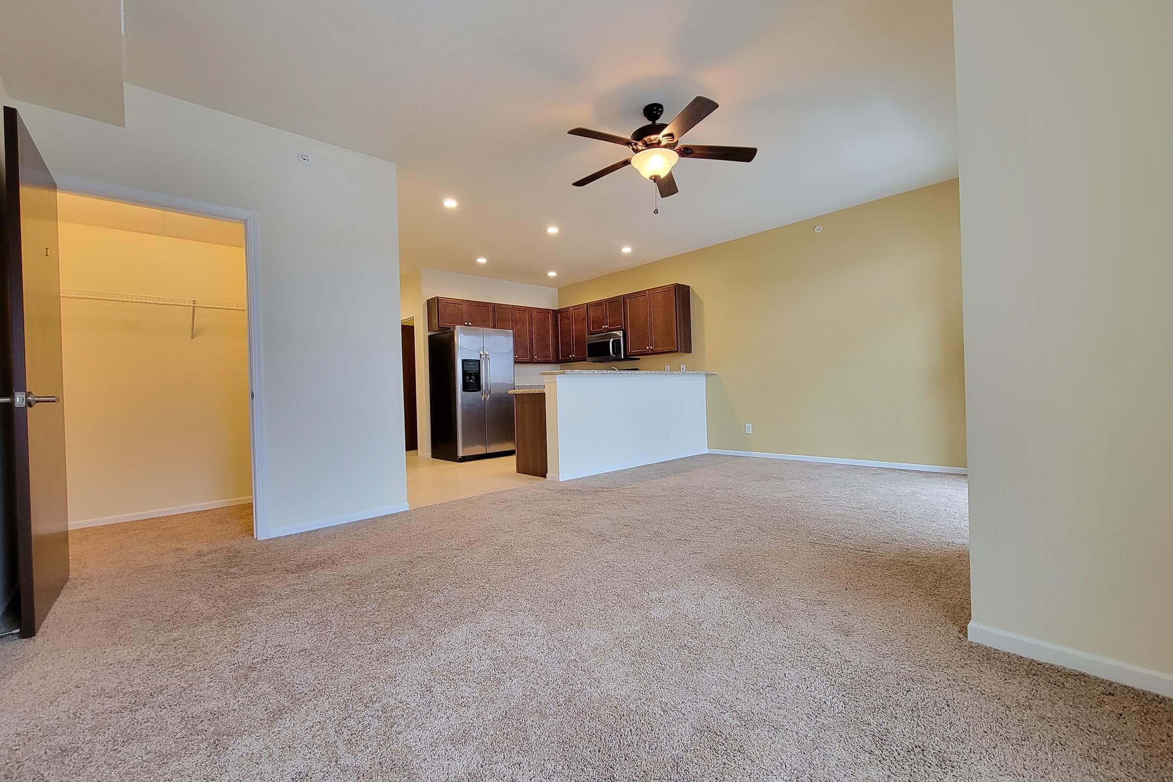 Spacious living area in a modern apartment featuring beige walls, carpeted flooring, and a ceiling fan. The area includes a kitchen with wooden cabinets and stainless steel appliances. A door leads to a closet, and there is ample natural light from the windows.