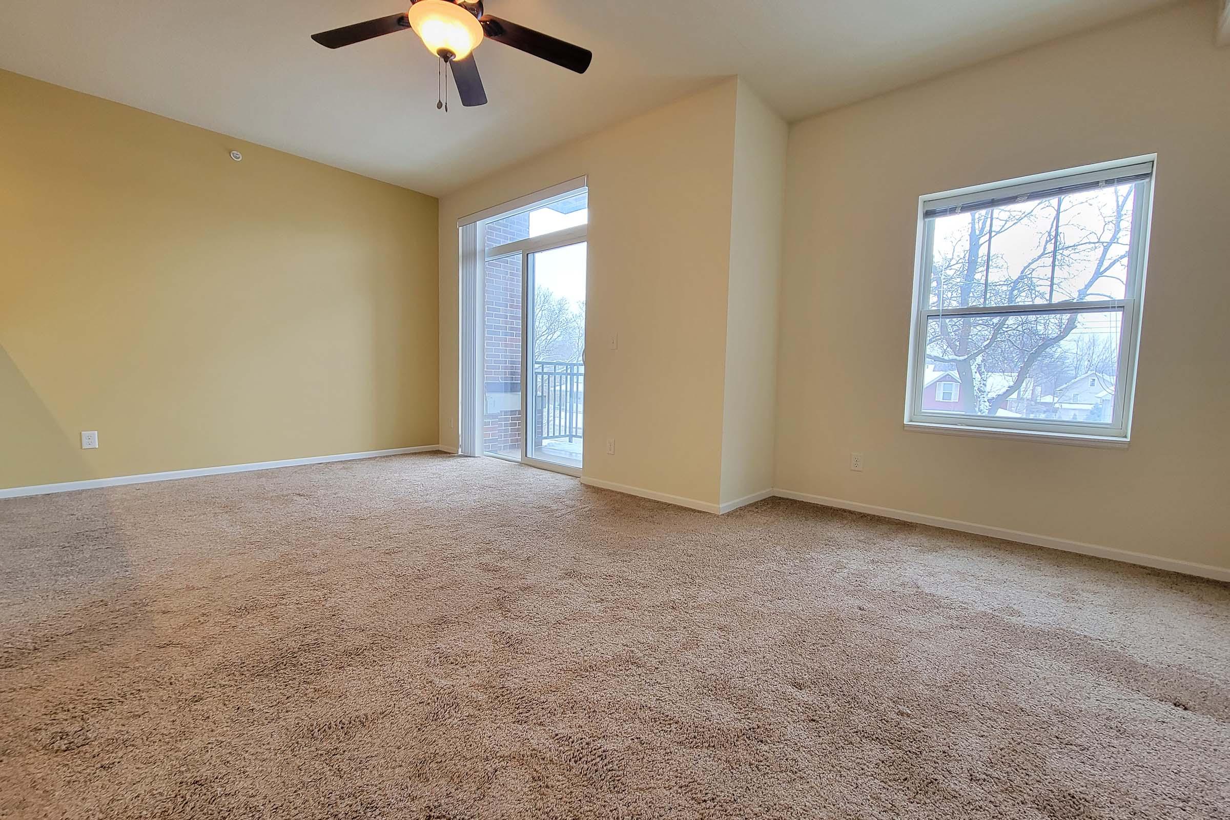 A spacious living room featuring light brown carpet, a ceiling fan, and a large window overlooking a balcony. The walls are painted a soft yellow, and there is ample natural light entering the room. The area is uncluttered and ideal for furniture arrangement.