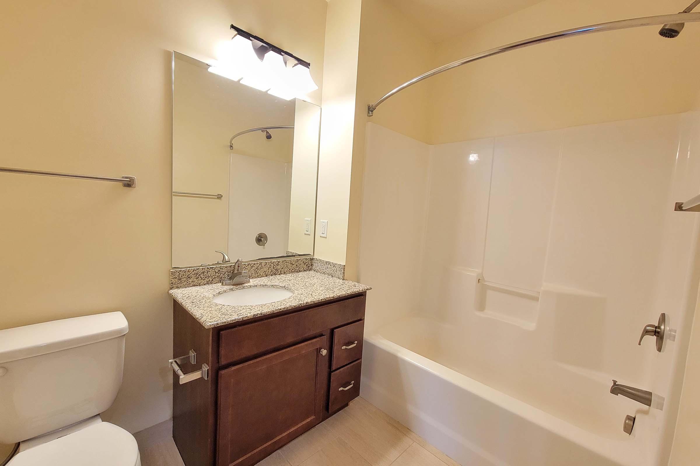 A clean and modern bathroom featuring a white bathtub with a curved shower rod, a single sink with a granite countertop, a wooden cabinet beneath, a large mirror above the sink, and a toilet. The walls are painted a soft yellow, creating a bright and inviting atmosphere.