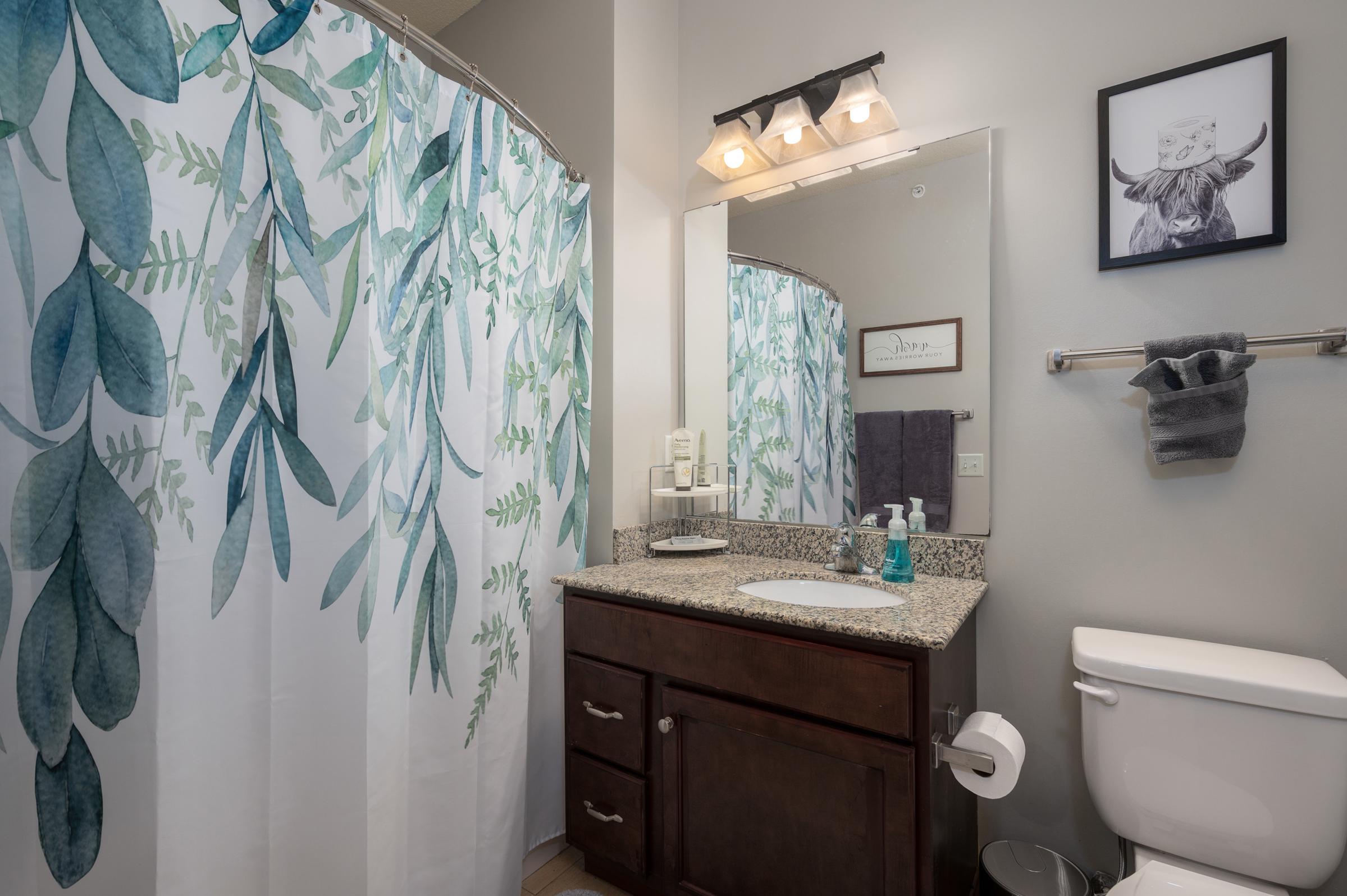 A modern bathroom featuring a light-colored wall, a large mirror above a dark wooden vanity with a granite countertop. A decorative shower curtain with leafy designs hangs in front of a bathtub. There is a toilet to the right, and a wall-mounted towel holder with a gray towel. A framed artwork is displayed above the vanity.
