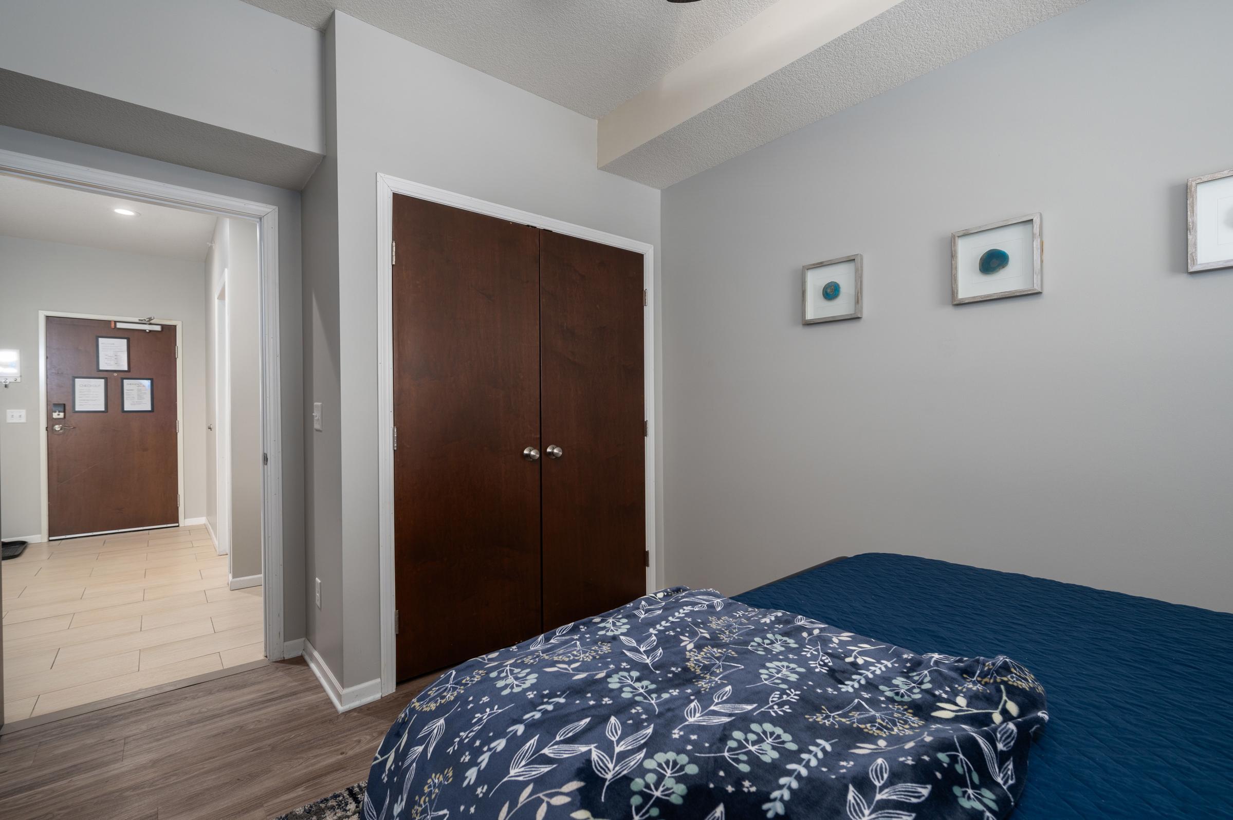 A cozy bedroom featuring a blue bedspread over a bed, with floral patterns. Two closed wooden doors are visible on the wall opposite the bed, and there are three framed circular artworks hanging on the gray wall. A hallway entrance can be seen in the background, and the flooring is light-colored tiles.