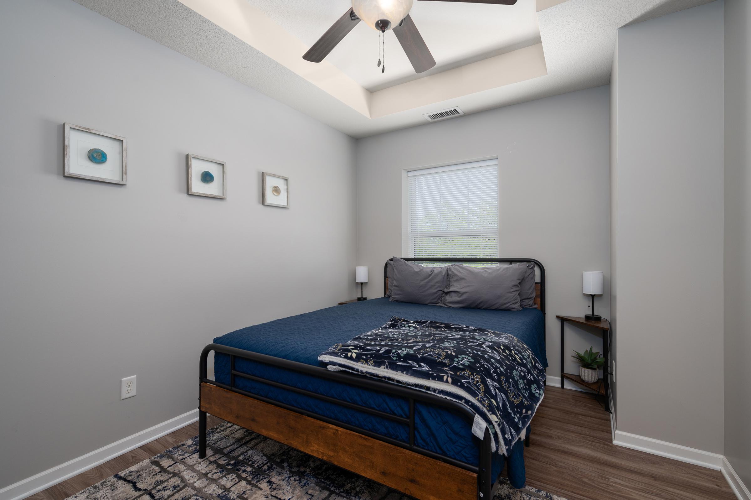 A cozy bedroom featuring a bed with blue bedding, a decorative throw, and a wooden frame. The room has light gray walls, a ceiling fan, and two bedside lamps. A window allows natural light, and there are three framed art pieces on the wall, along with a small plant in the corner. The flooring is laminate.
