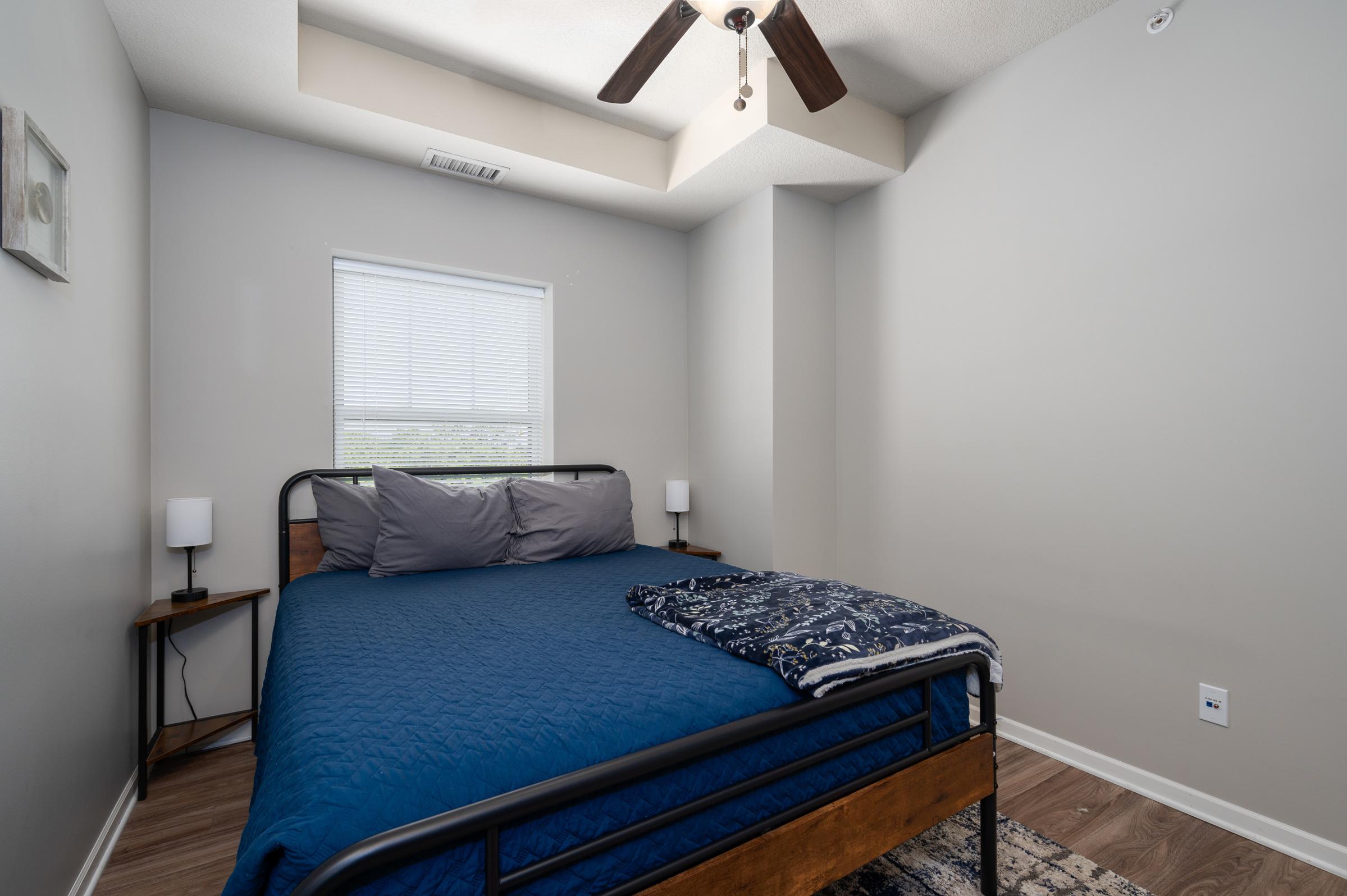 A cozy bedroom featuring a queen-sized bed with blue bedding, two bedside lamps on wooden nightstands, and a small area rug. The window allows natural light, and the room has neutral-colored walls and a ceiling fan.