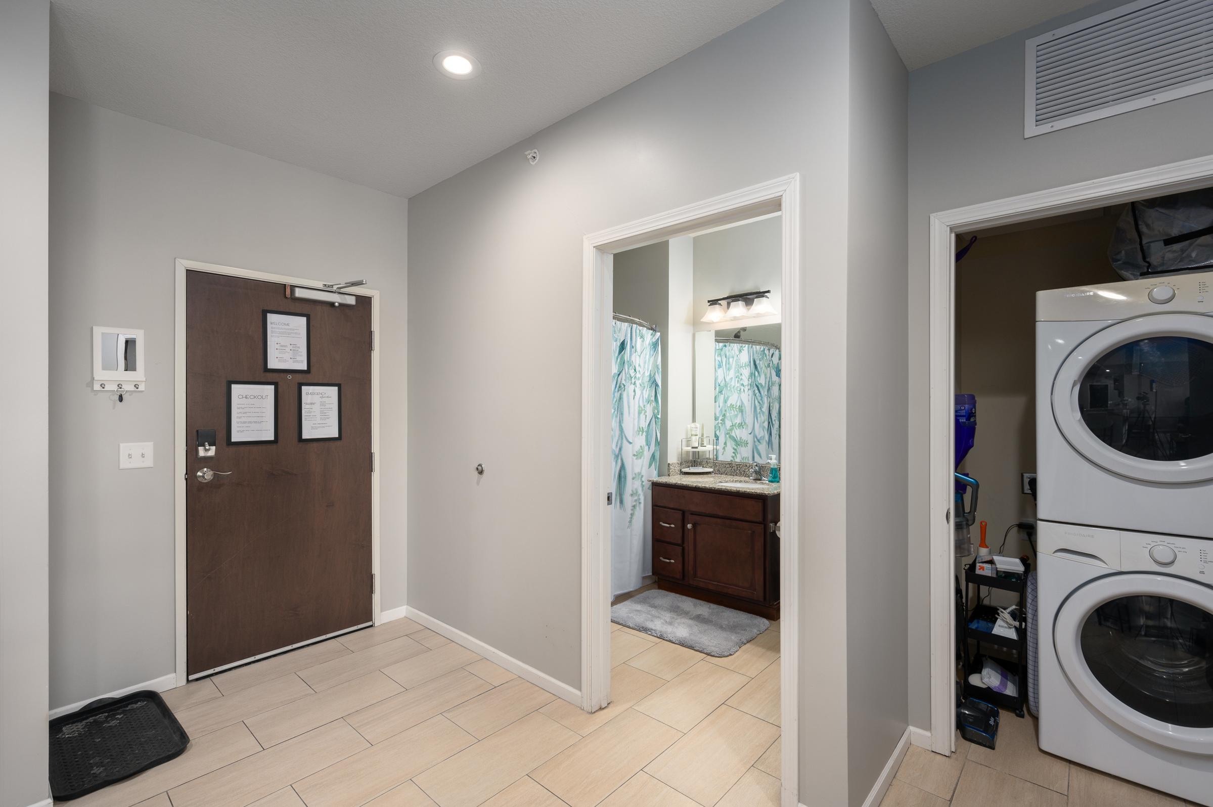 A well-lit entryway featuring a wooden door on the left, leading to a bathroom with a vanity and blue patterned shower curtain. To the right, a laundry area with a stacked washer and dryer. Light-colored walls and tiled flooring create a modern and inviting atmosphere.