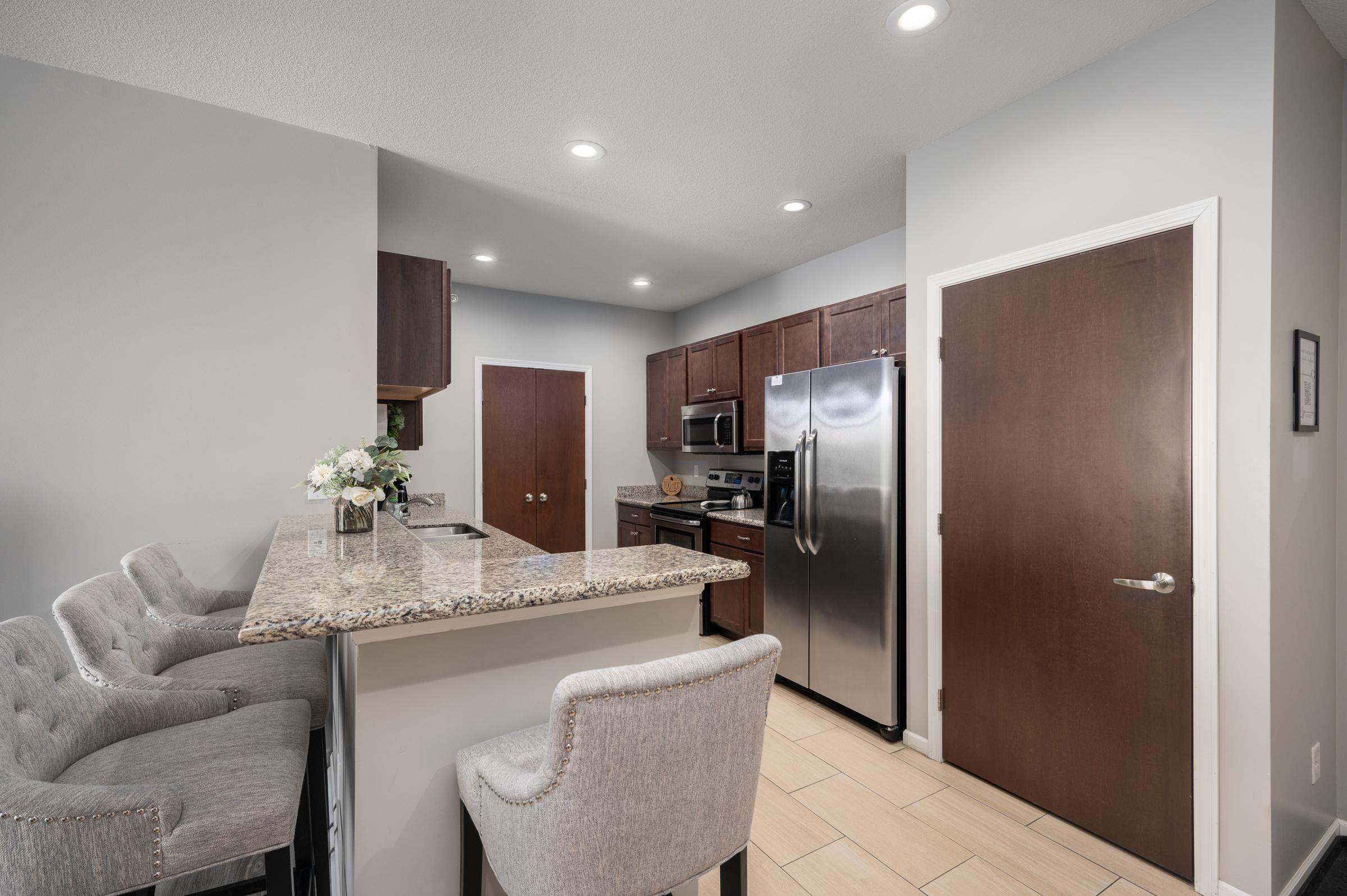 Modern kitchen with granite countertops, wood cabinets, stainless steel appliances, and bar seating. The space features soft gray walls, recessed lighting, and a closed door leading to another room, creating a stylish and functional atmosphere.