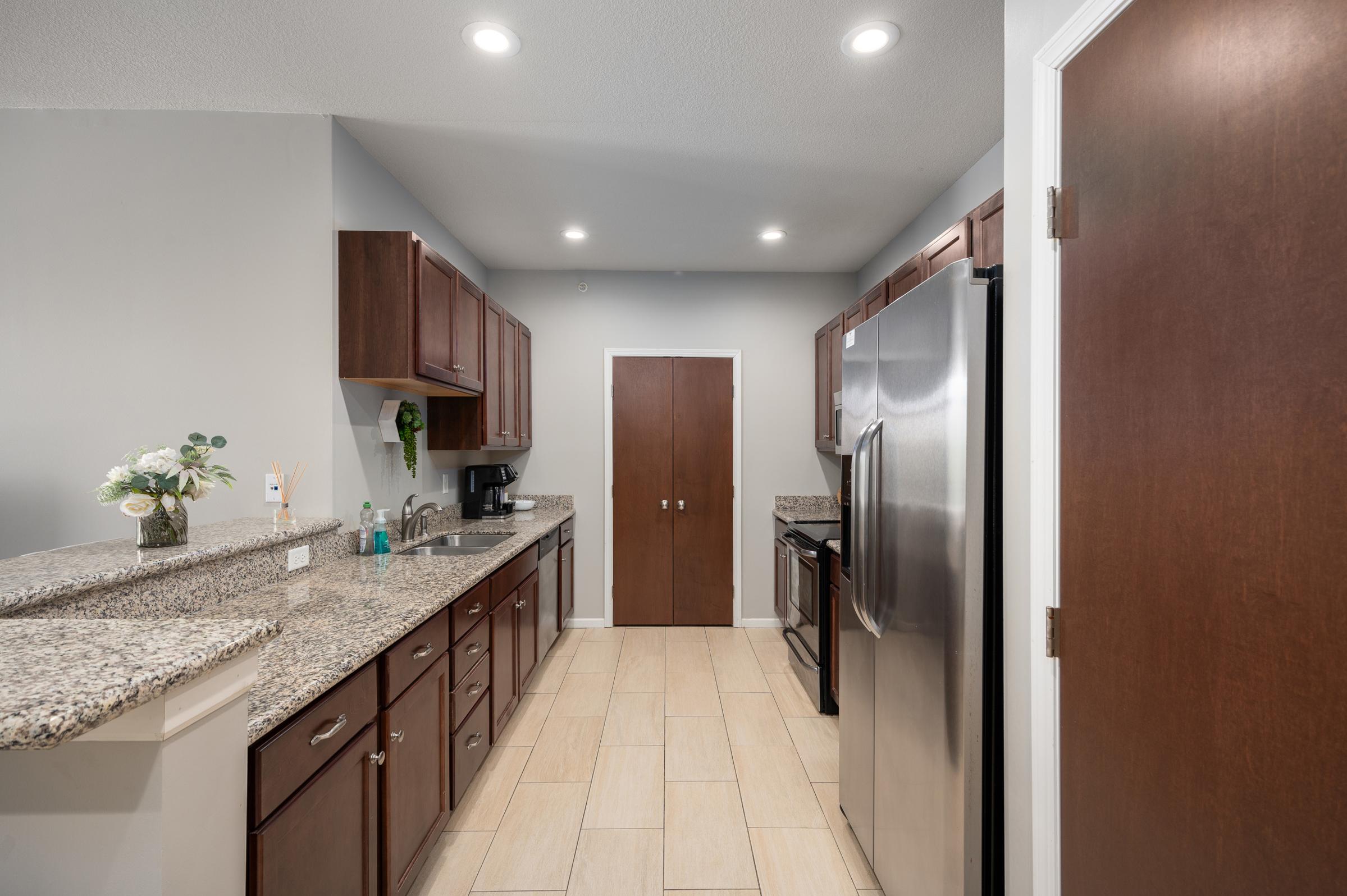 Modern kitchen featuring dark wood cabinetry, granite countertops, stainless steel appliances, and a light-colored tile floor. There is a door at the end of the kitchen, and the counter is decorated with a small plant and kitchen essentials. Bright overhead lights illuminate the space.
