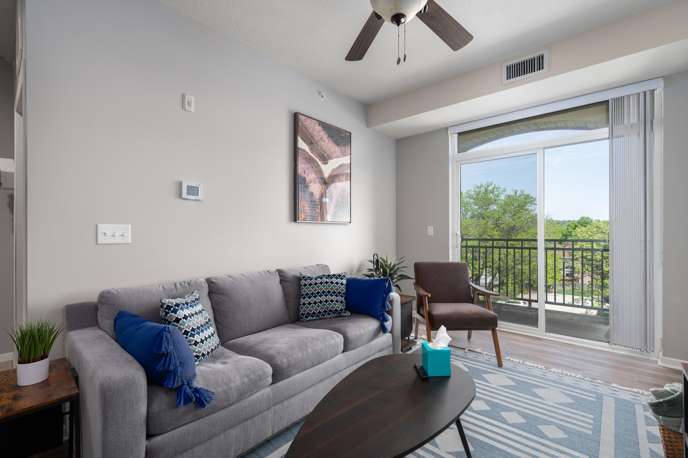 A modern living room featuring a gray sofa with blue and white decorative pillows, a wooden coffee table, and a potted plant. There’s a window with a view of greenery, a ceiling fan, and artwork on the wall. A cozy armchair next to the sofa adds to the inviting atmosphere.