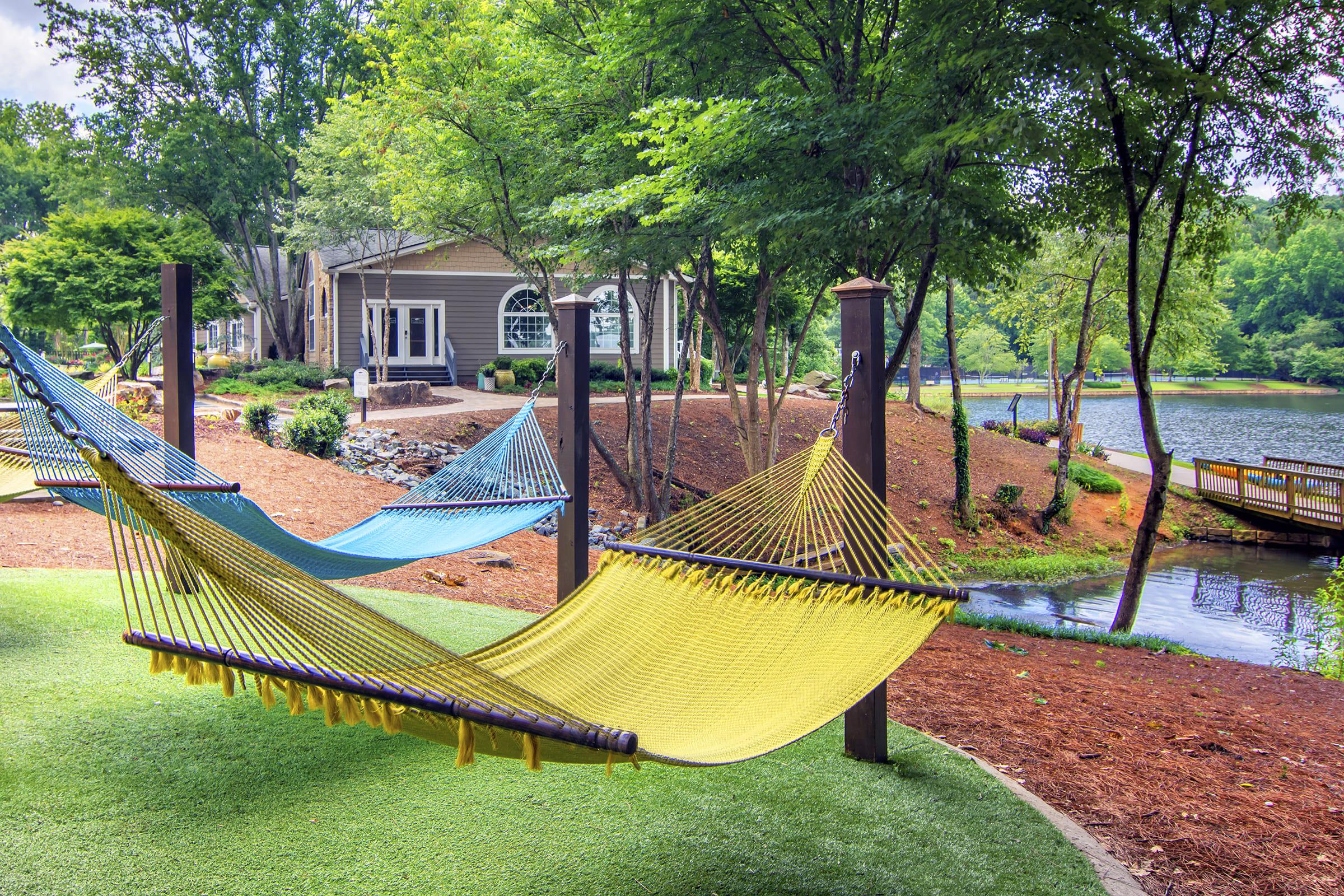 Two colorful hammocks, one yellow and one blue, are suspended between wooden posts in a peaceful outdoor setting. Lush green trees surround the area, and a calm lake is visible in the background, reflecting the natural landscape. A cozy house with large windows is nearby, creating a serene atmosphere.