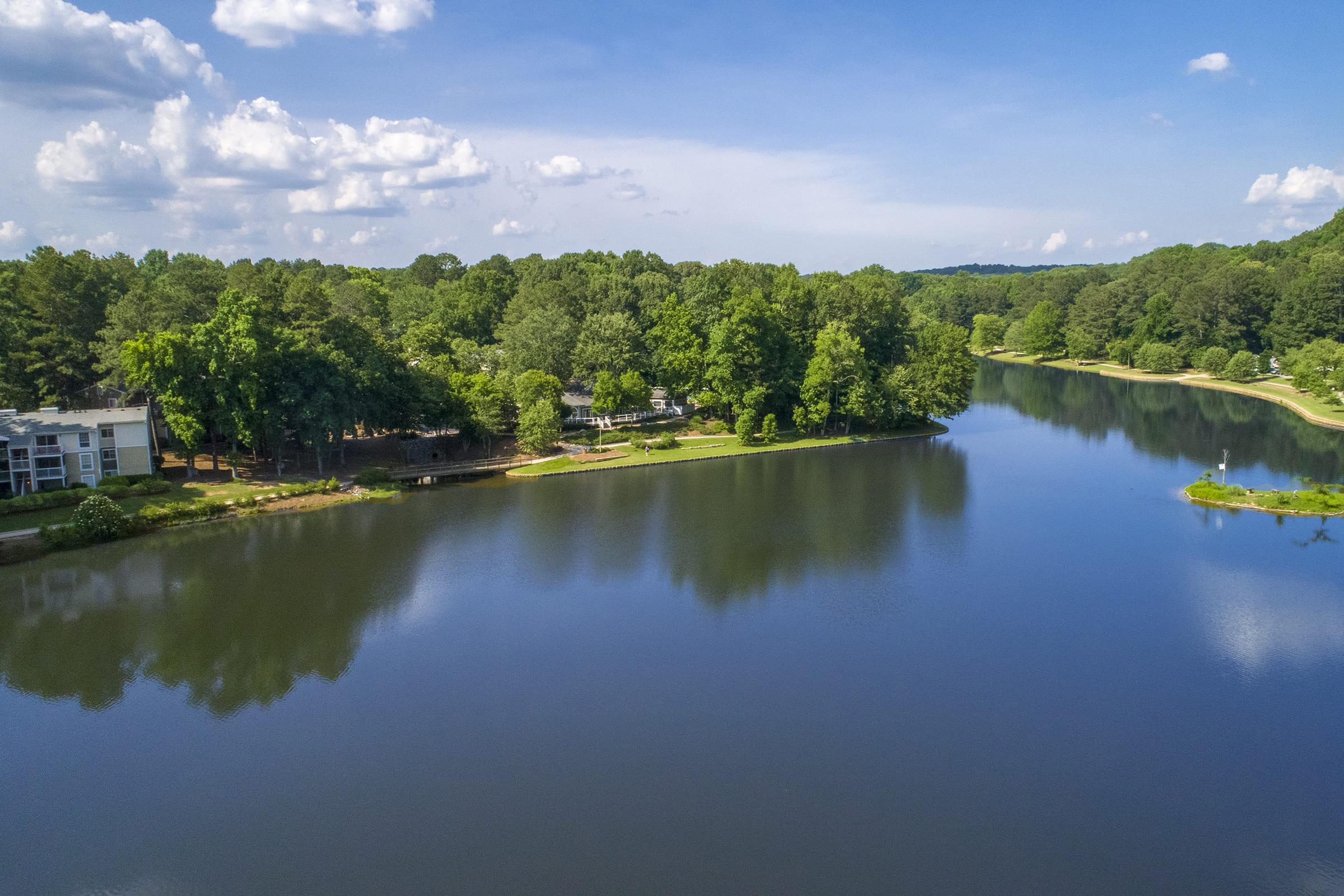 A serene view of a calm lake surrounded by lush green trees under a blue sky with scattered clouds. On one side, there are residential buildings near the water's edge, creating a peaceful natural setting ideal for relaxation and outdoor activities.
