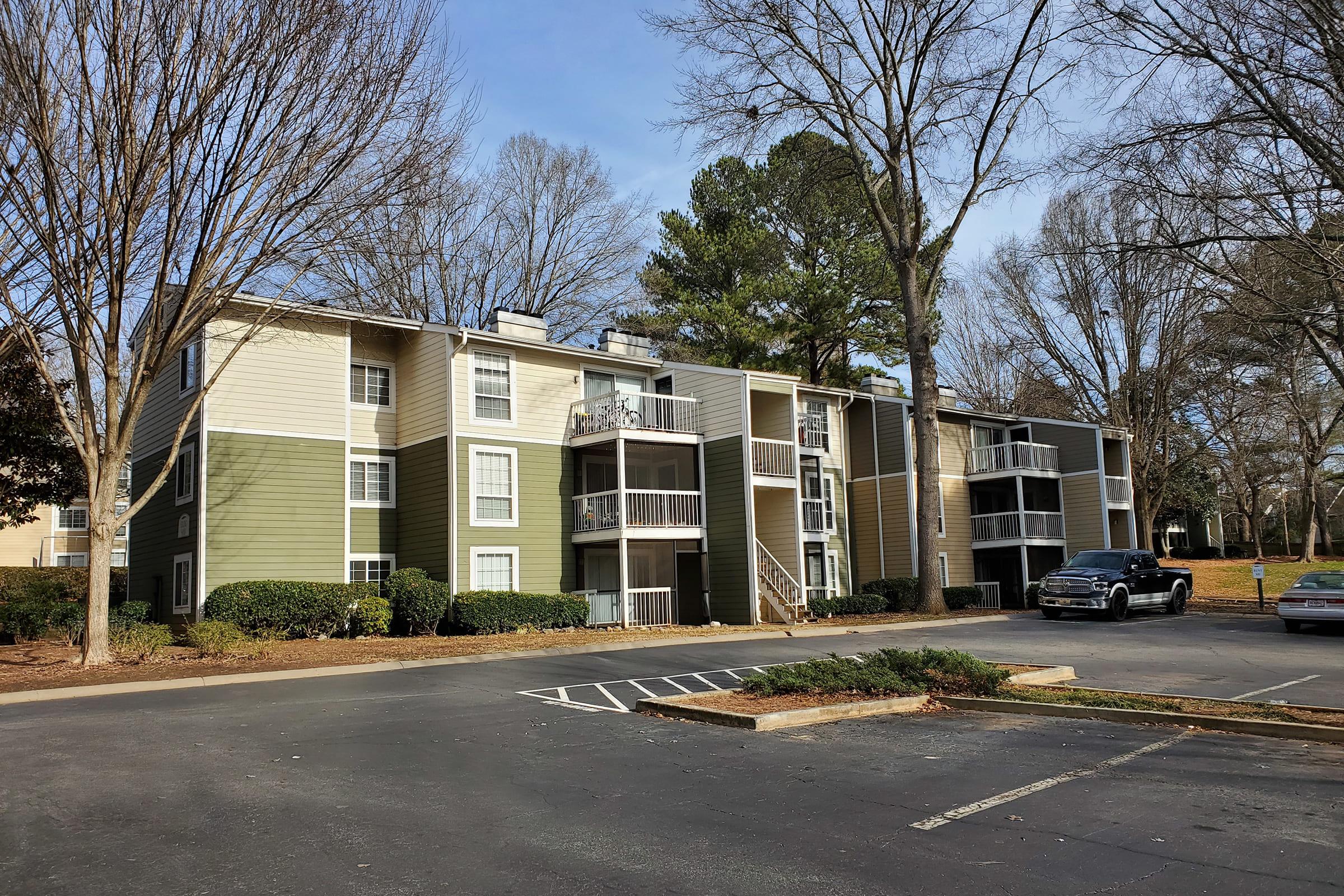A multi-unit apartment building featuring a combination of green and beige siding. The building has several balconies, surrounded by trees without leaves, and a parking area in the foreground with a few empty spaces. It's a clear day with blue skies.