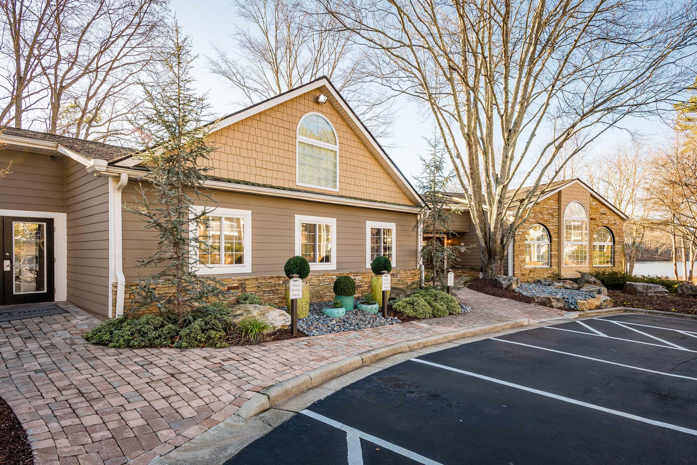 A cozy building with a sloped roof and large windows, surrounded by well-maintained landscaping featuring small trees, shrubs, and decorative stones. The approach to the entrance includes a brick pathway and a parking area. The setting appears serene, with a clear sky and natural surroundings.