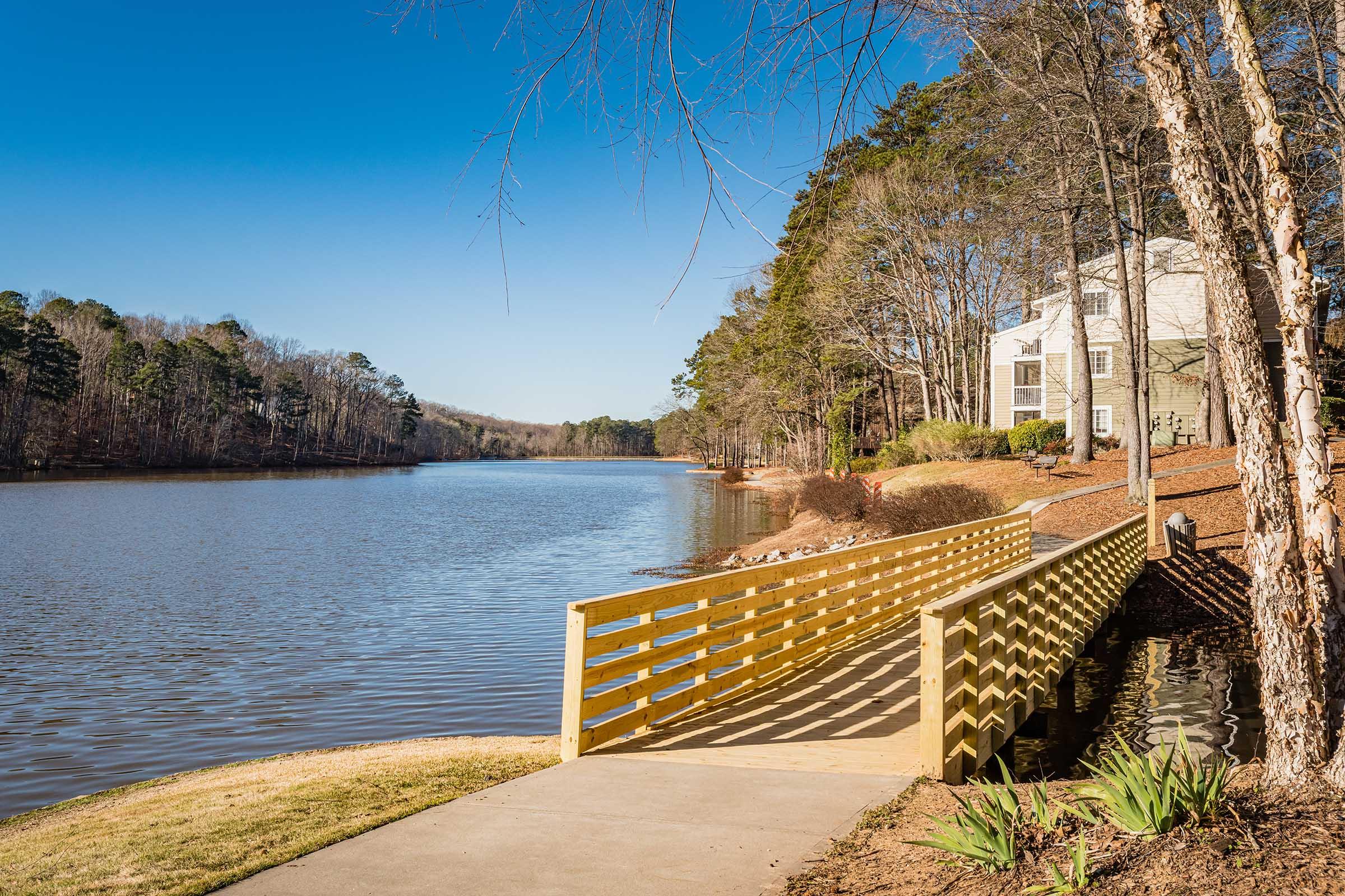 A serene lakeside scene featuring a wooden walkway leading to the water, bordered by grassy areas and trees. On the right, there are modern buildings tucked among the greenery. The sky is clear and blue, reflecting a calm atmosphere on the lake's surface.