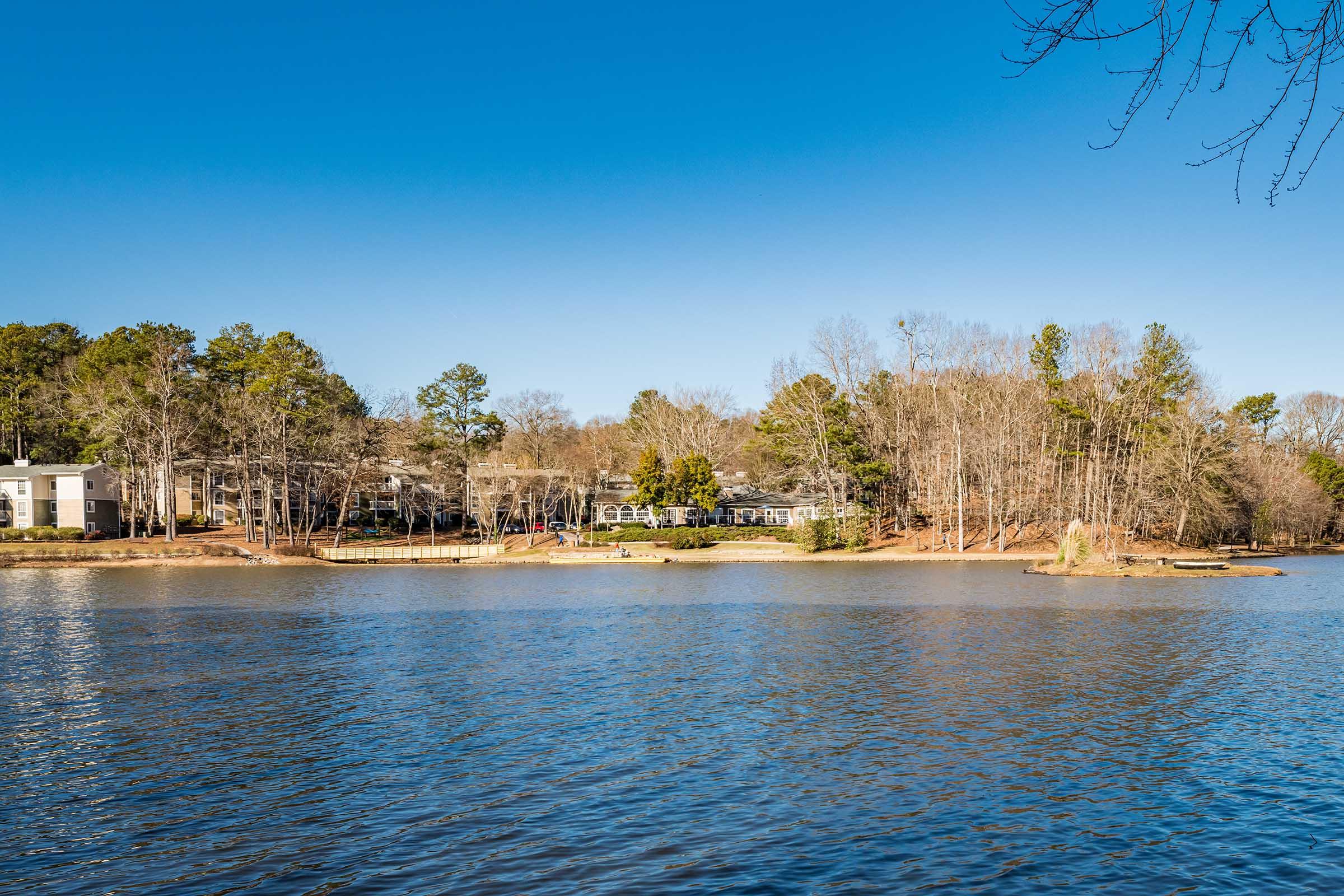 A tranquil lake scene with clear blue skies, featuring a shoreline lined with bare trees and a few houses in the background. The water reflects the calm surroundings, creating a peaceful atmosphere. A small island with trees is visible in the distance, enhancing the natural beauty of the landscape.