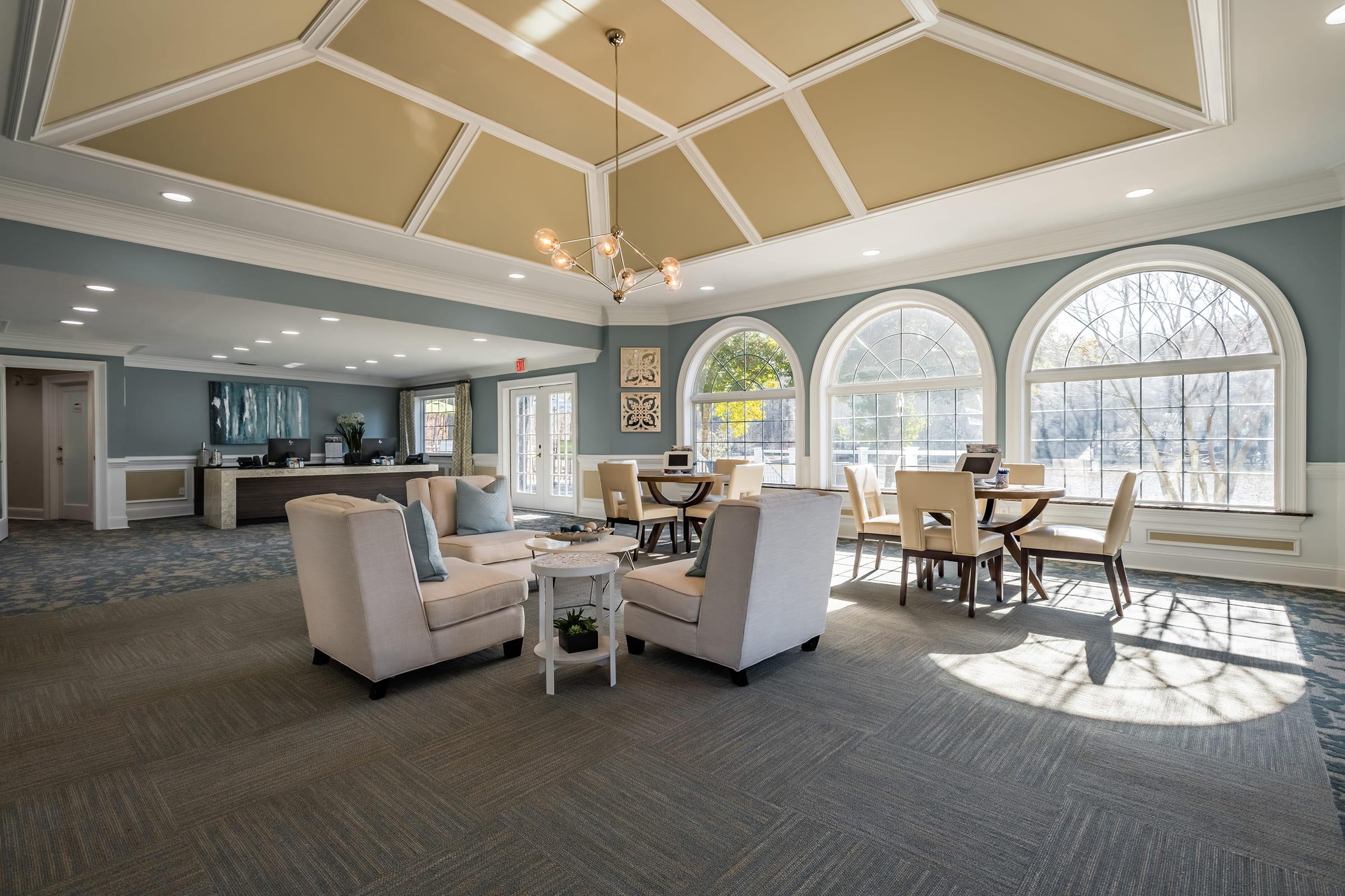 Bright and airy lounge area featuring large arched windows, a cozy seating arrangement with light-colored chairs, and a small table. Sunlight filters in, creating a warm atmosphere. In the background, there is a modern kitchenette and dining space with wooden tables.