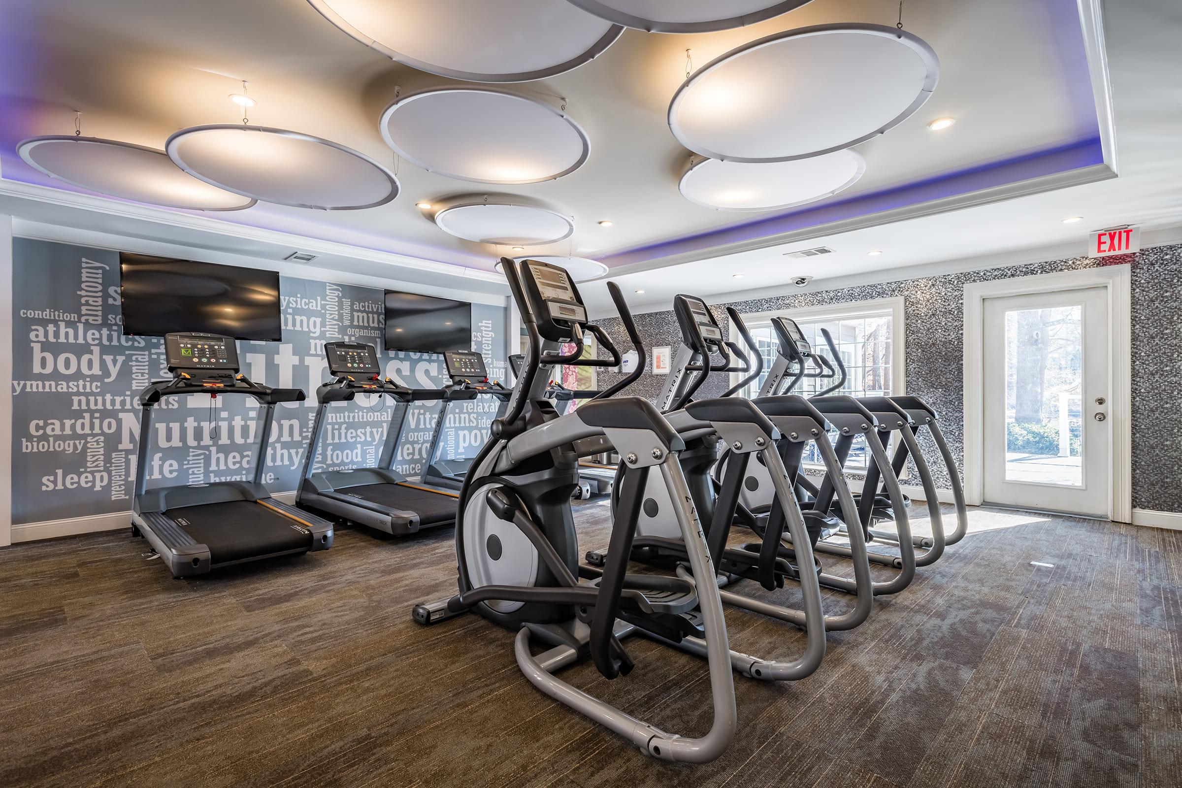 A modern fitness center featuring several cardio machines, including treadmills and ellipticals. The room has a stylish design with circular ceiling lights and motivational words on the walls, creating an inviting atmosphere for exercise. A door leads to the outside, emphasizing accessibility.