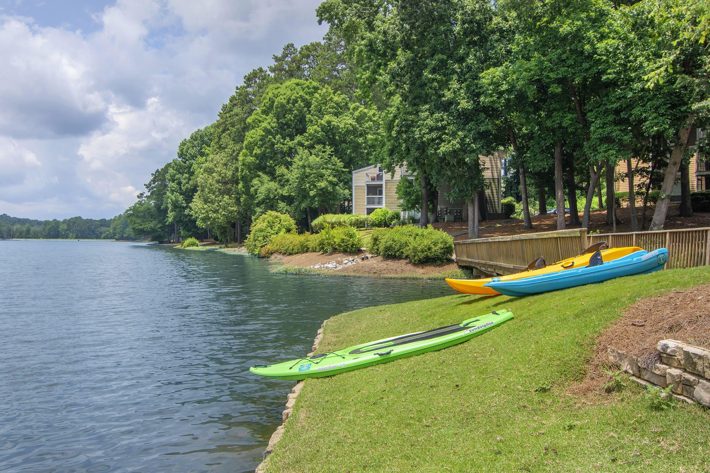 Two kayaks, one green and one yellow, are resting on a grassy bank by a calm lake. In the background, trees provide shade, and there are residential buildings nestled among the greenery. The sky is partly cloudy, reflecting a peaceful outdoor setting ideal for kayaking and enjoying nature.