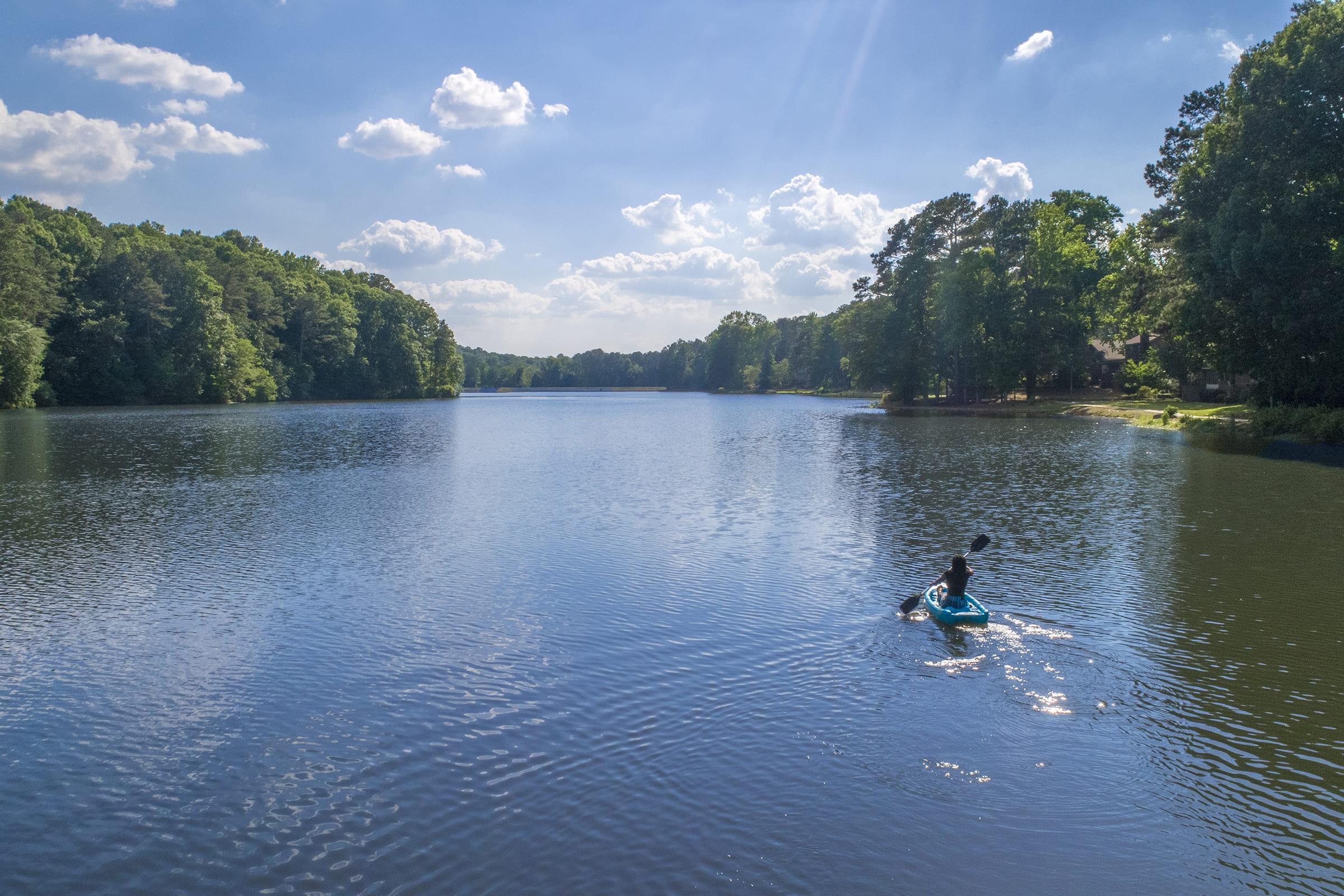 A calm lake surrounded by lush green trees under a bright blue sky with fluffy white clouds. A person is kayaking in the foreground, creating ripples in the water. The scene conveys tranquility and natural beauty, ideal for outdoor activities.