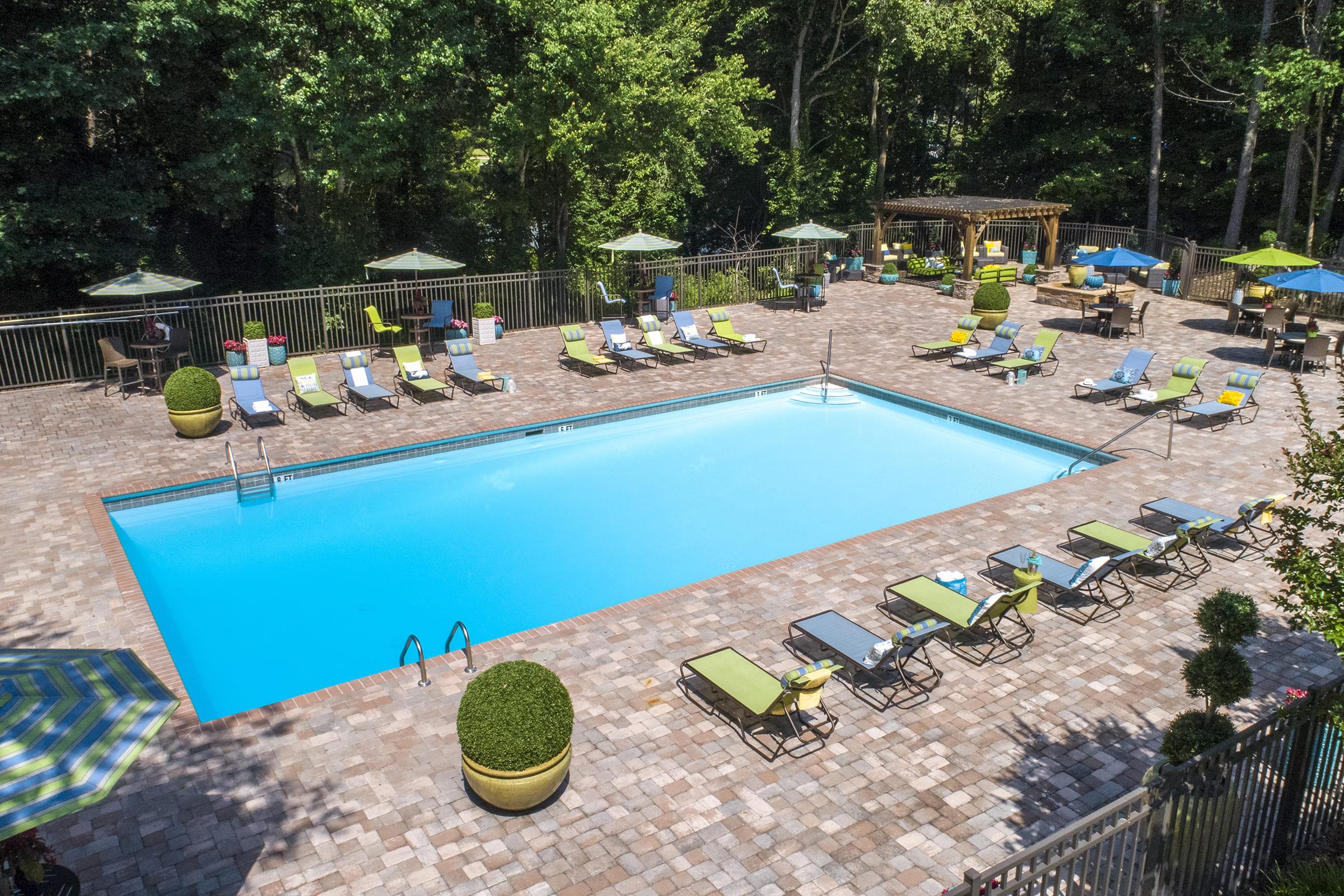 A sunny outdoor pool surrounded by lounge chairs and umbrellas. The area features a well-maintained brick patio with greenery and seating areas. Trees provide shade, creating a relaxing atmosphere for leisure and recreational activities.