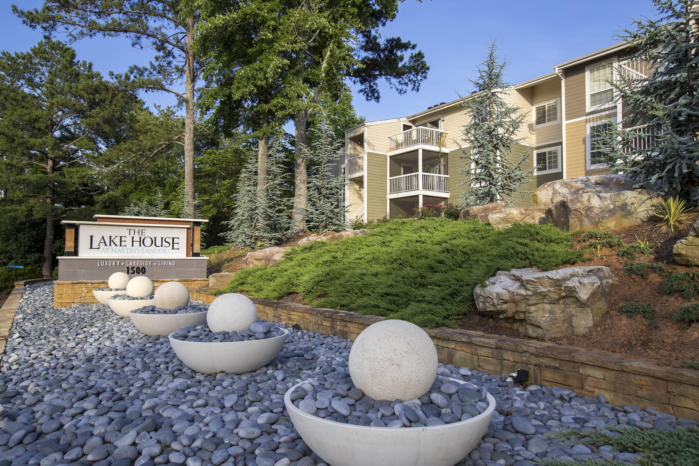 The image shows the entrance to "The Lake House" apartment complex, featuring a sign with the name and address. Neatly landscaped grounds include large spherical decorations in stone planters and greenery, with several tall trees and a building visible in the background.