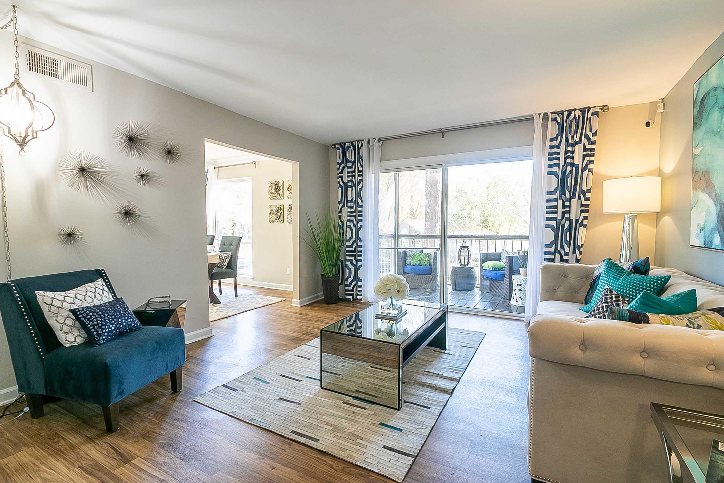 A modern living room featuring a blue accent chair, a beige sofa with colorful pillows, a glass coffee table, and a decorative rug. Large windows provide natural light, and there are elegant curtains with a bold pattern. The space includes decorative wall art and plants, creating a bright and inviting atmosphere.