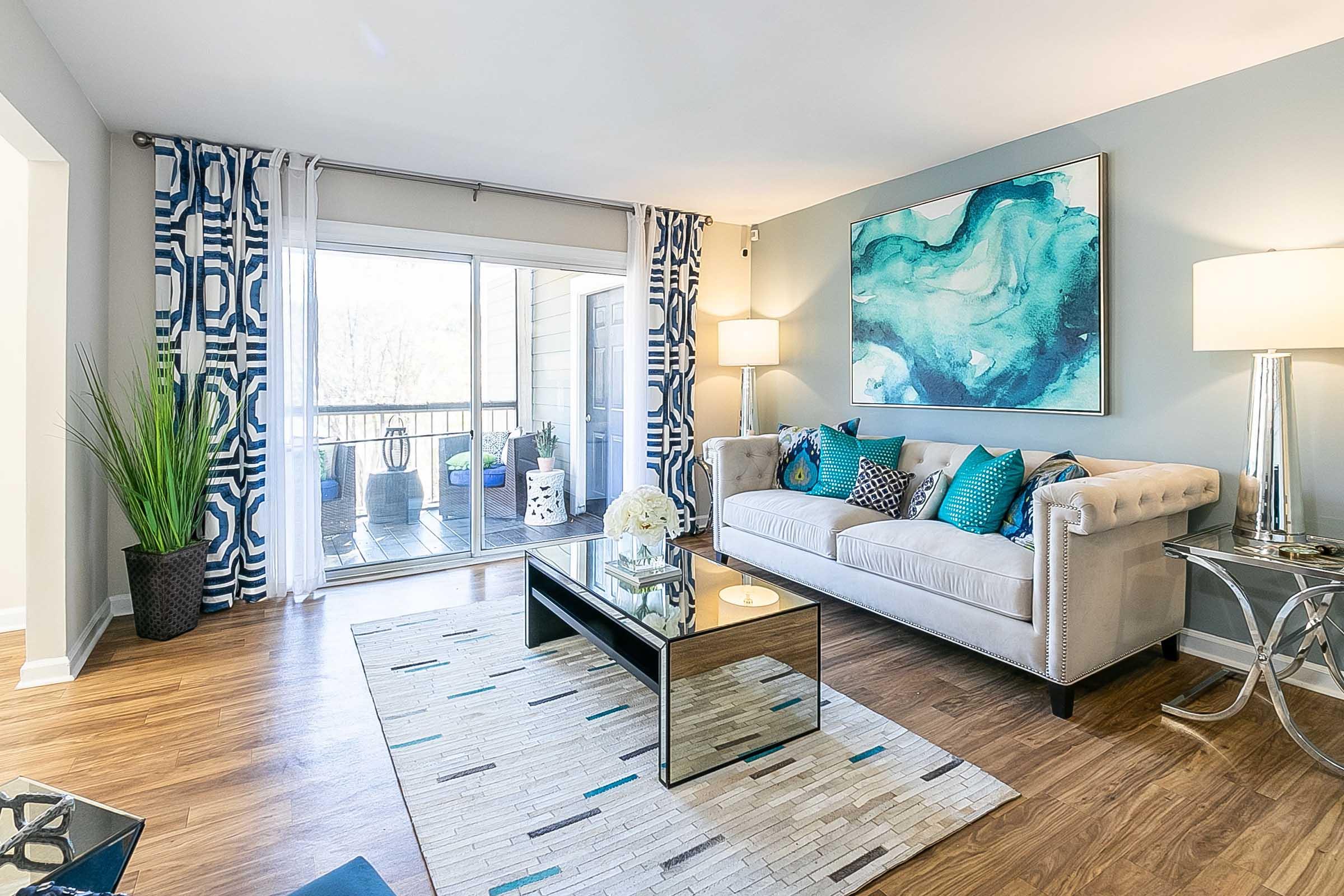 A modern living room featuring a light-colored tufted sofa adorned with teal and patterned cushions. The room has large windows with sheer curtains, letting in natural light. A sleek coffee table sits on a decorative area rug, and a potted plant adds a touch of greenery. The walls are painted in soft tones, complemented by blue and silver decor.