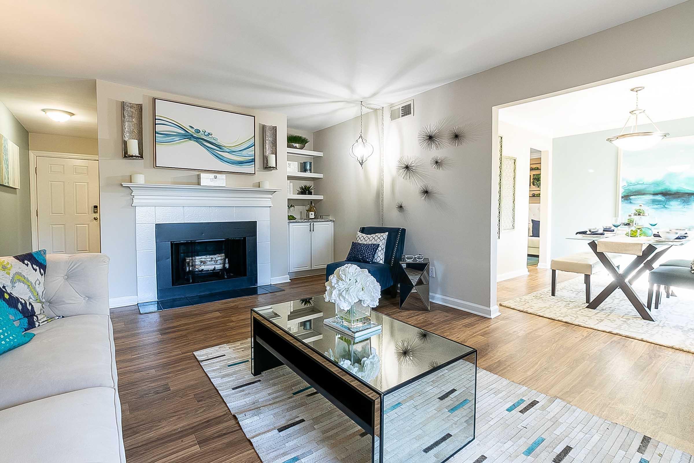 A modern living room featuring a cozy fireplace, decorative shelves, and a glass coffee table. There are comfortable seating options, including a light sofa and stylish accents, with a piece of artwork above the fireplace. Natural light flows in, enhancing the inviting atmosphere of the space.