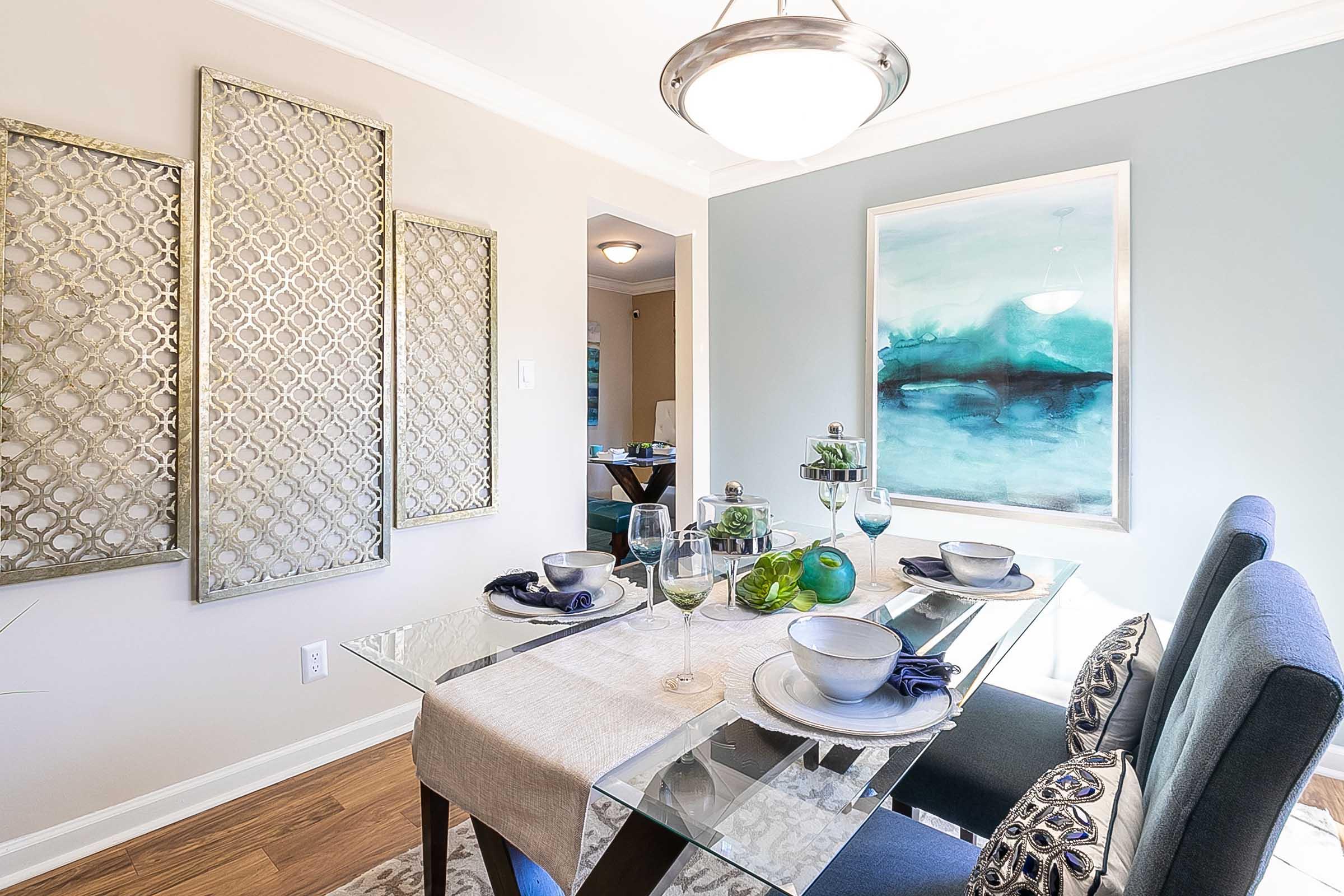 A modern dining area featuring a glass table set for a meal with blue and white dishware, a table runner, and decorative items. The walls are adorned with stylish artwork and metallic wall décor, creating a contemporary and inviting atmosphere, complemented by elegant lighting fixtures.
