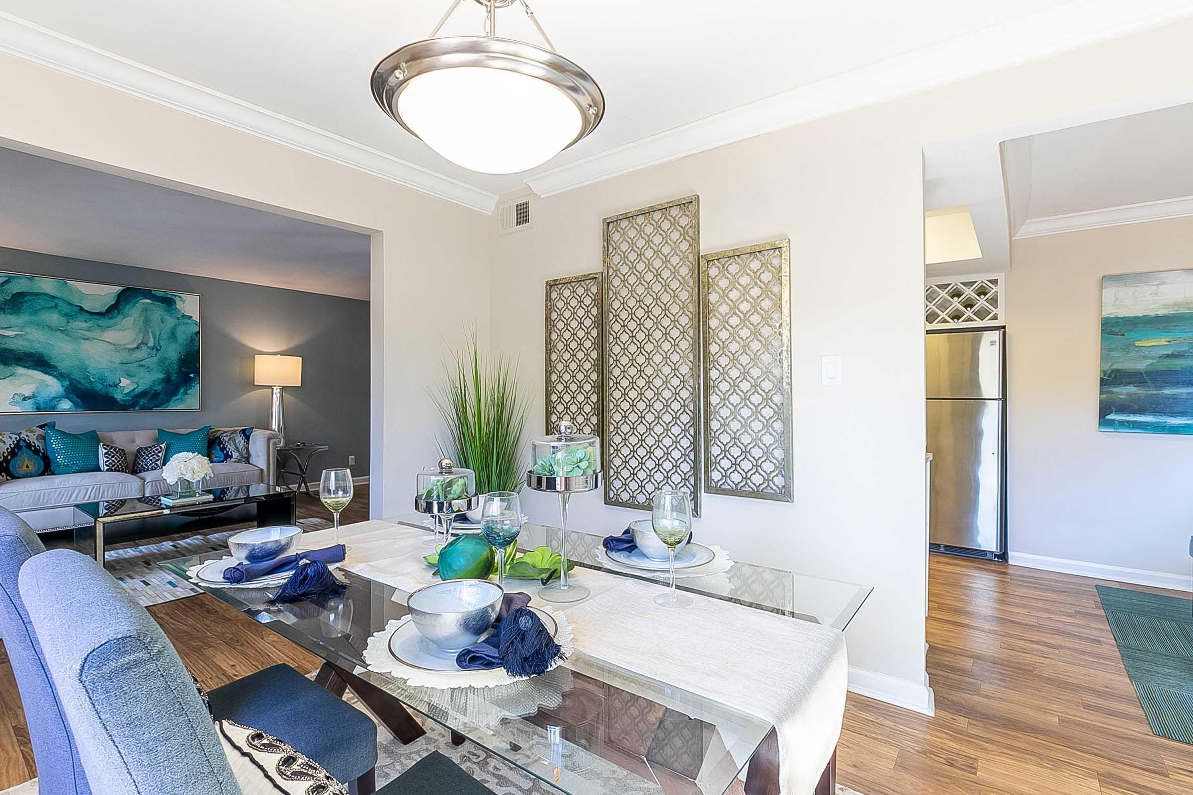 Elegant dining area featuring a glass table set with decorative bowls and glassware. The space showcases modern décor with a neutral color palette, a large light fixture, and artwork on the walls. A living area is visible in the background, adding to the inviting atmosphere.