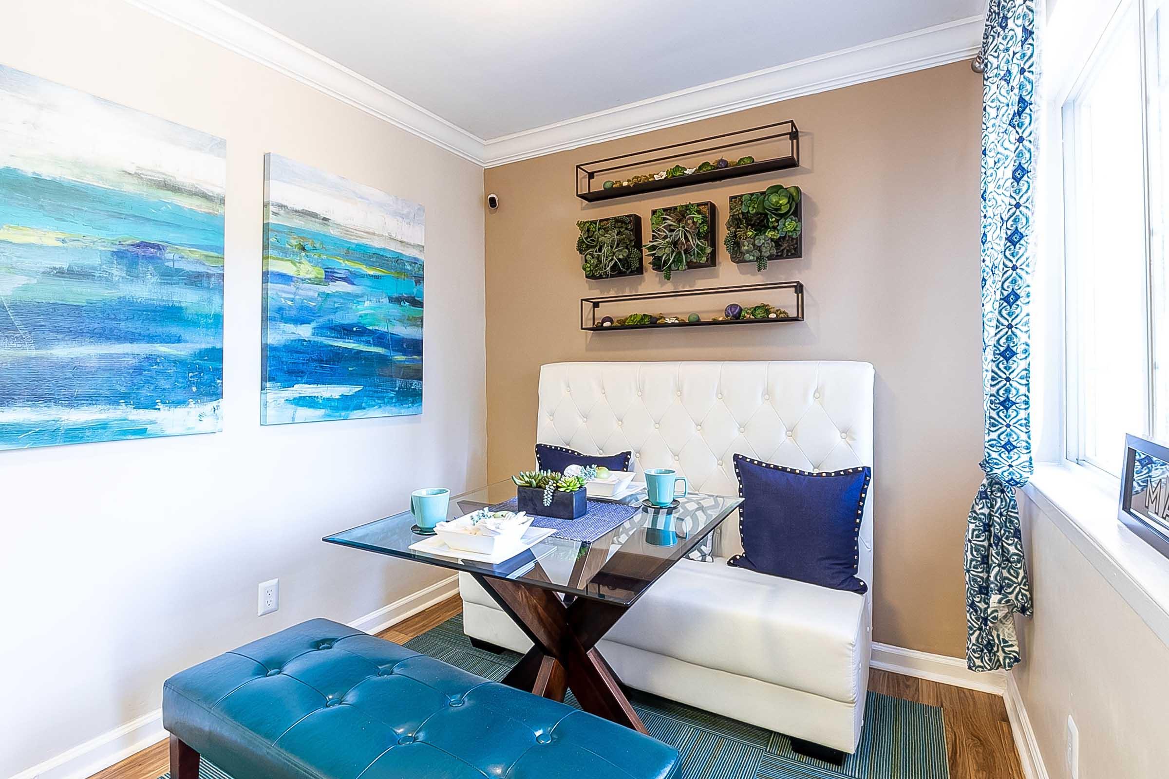 Cozy dining nook featuring a glass table with a dark wooden base, surrounded by a white tufted bench and blue cushions. The walls are decorated with abstract blue artwork and a vertical garden shelf with greenery. Natural light filters in through a window with patterned curtains.