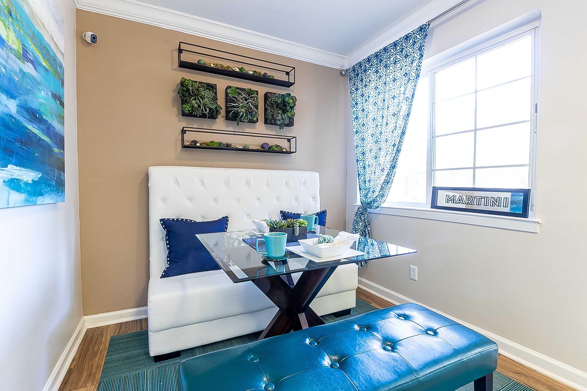 A cozy room featuring a white tufted loveseat with blue accent pillows, a glass dining table, and a teal ottoman. There are decorative shelves with plants on the wall and a window with patterned blue drapes. A sign reading "MARTINI" is displayed on the window ledge.