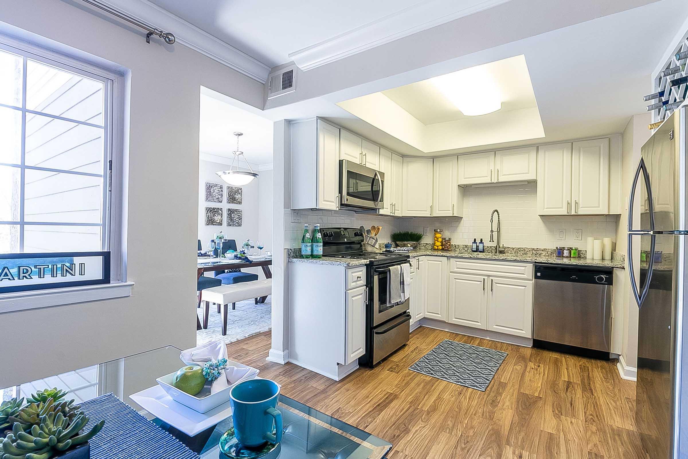 Modern kitchen featuring white cabinetry, stainless steel appliances, and a granite countertop. Natural light floods in through a window. A dining area is visible in the background, with a table set for four. Decor includes green plants and kitchen accessories, creating a clean and inviting atmosphere.