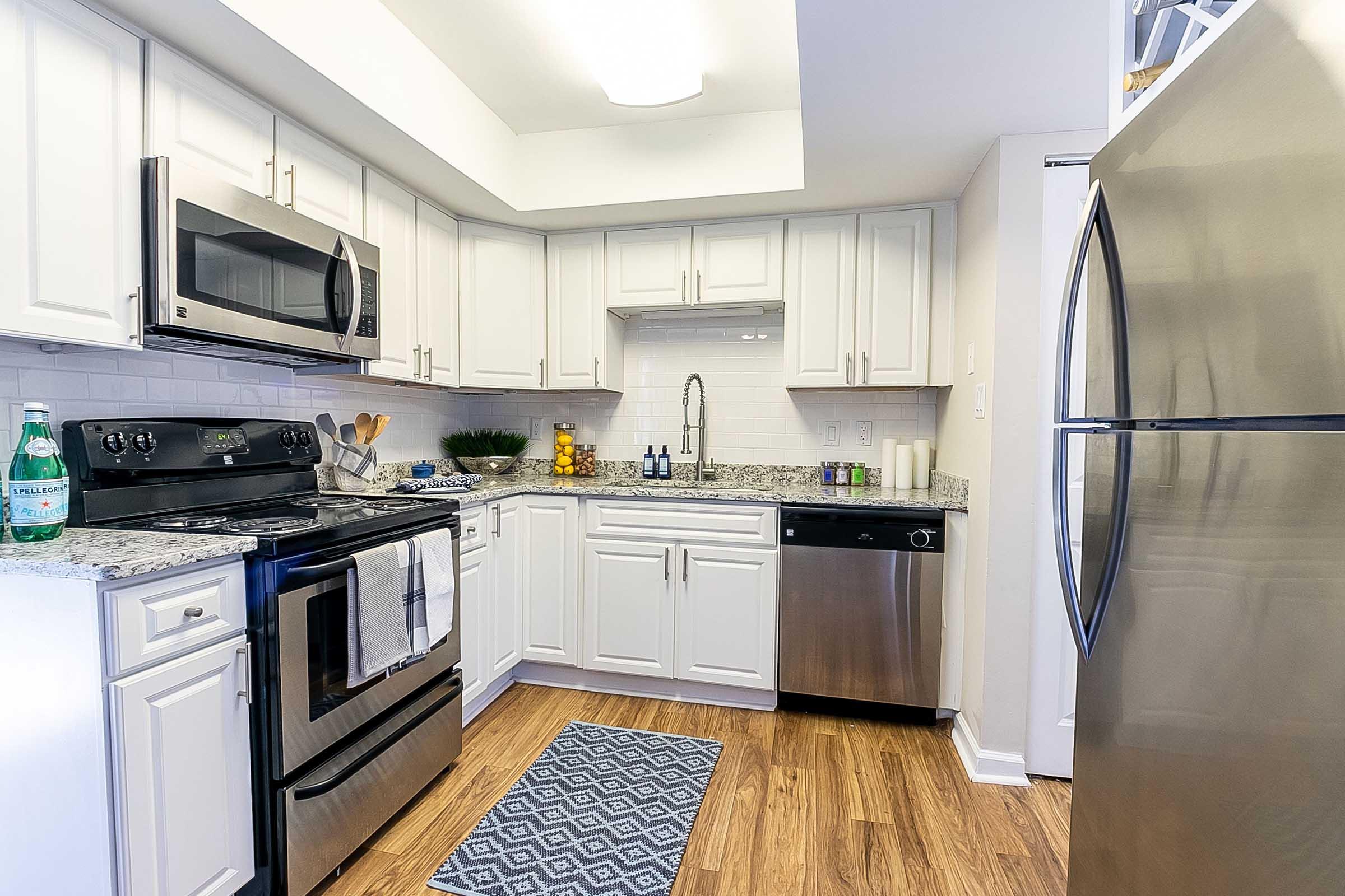 Modern kitchen featuring stainless steel appliances, including a microwave, oven, and dishwasher. The white cabinetry is complemented by a granite countertop and white backsplash. A rug adds warmth to the wooden floor. Decorative items, including fruit, are arranged neatly on the counter.